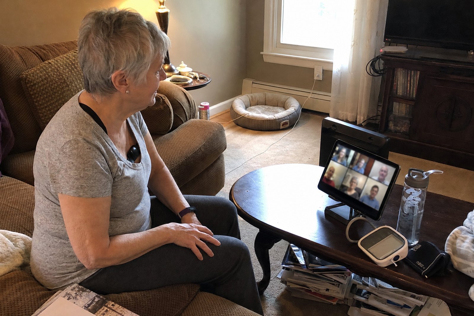 A woman around seventy on her own sofa wearing a chest-strap heart rate monitor and a wrist band, following a small video-call grid on a tablet propped on the coffee table beside a blood pressure cuff.