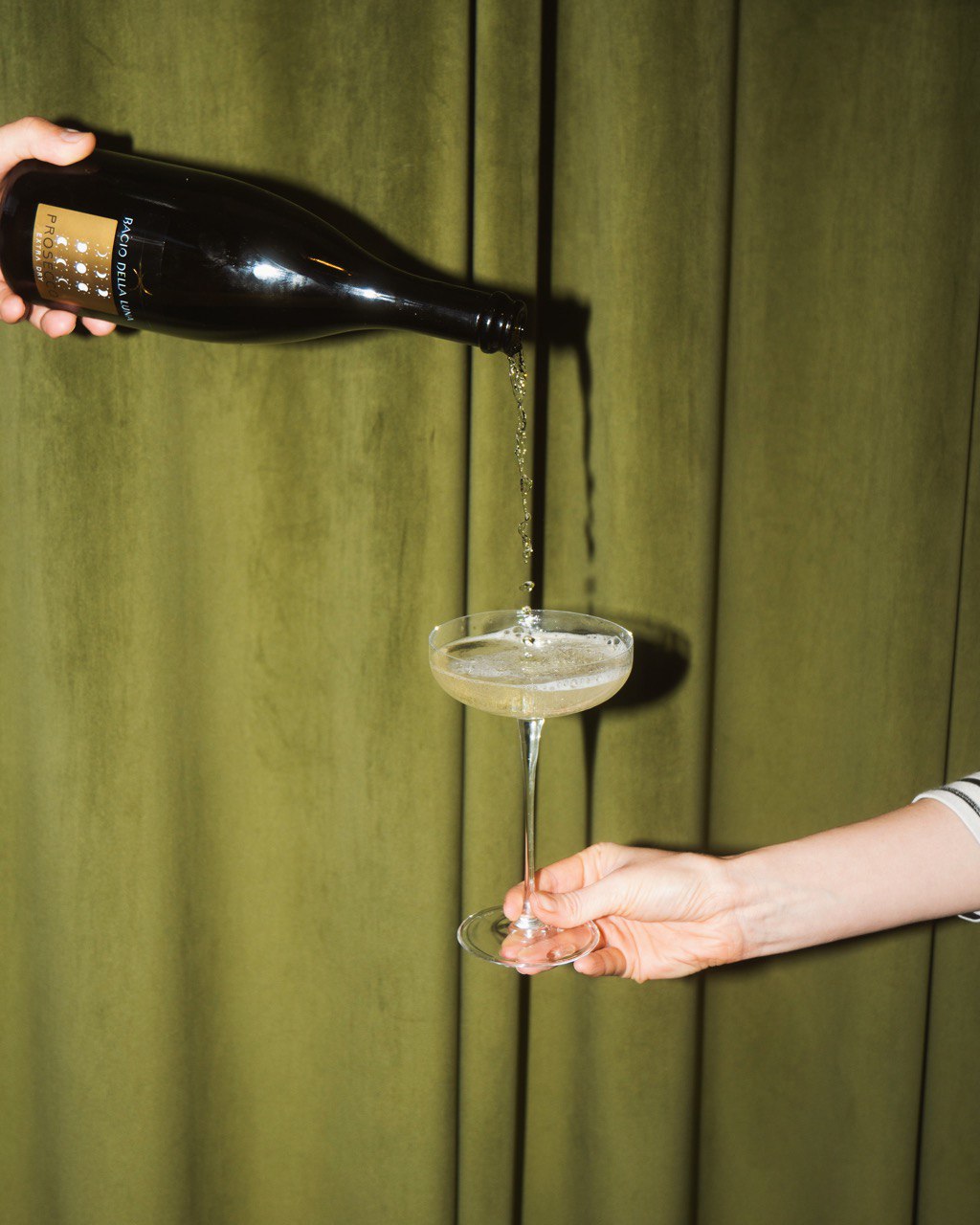 A bottle of sparkling wine is being poured into a coupe glass against a backdrop of olive-green curtains, featuring elegant bubbles forming as the liquid fills the glass.