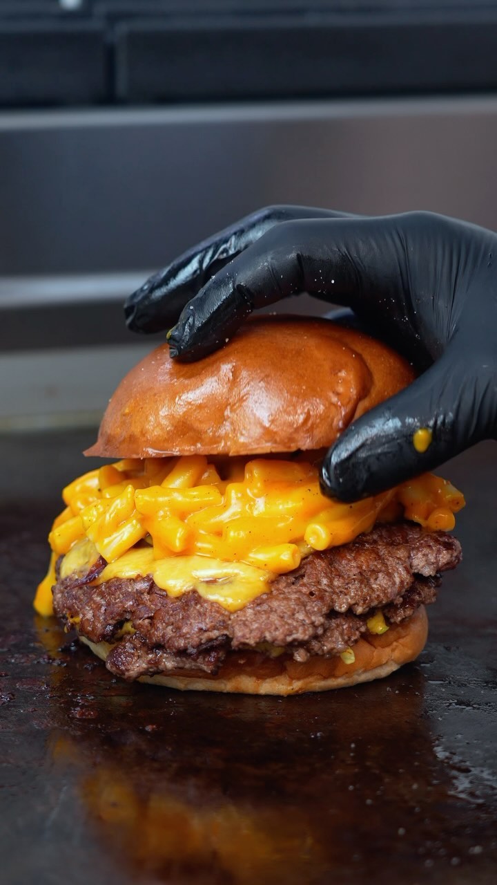 A gourmet burger topped with melted cheese, held with a gloved hand, on a dark countertop.