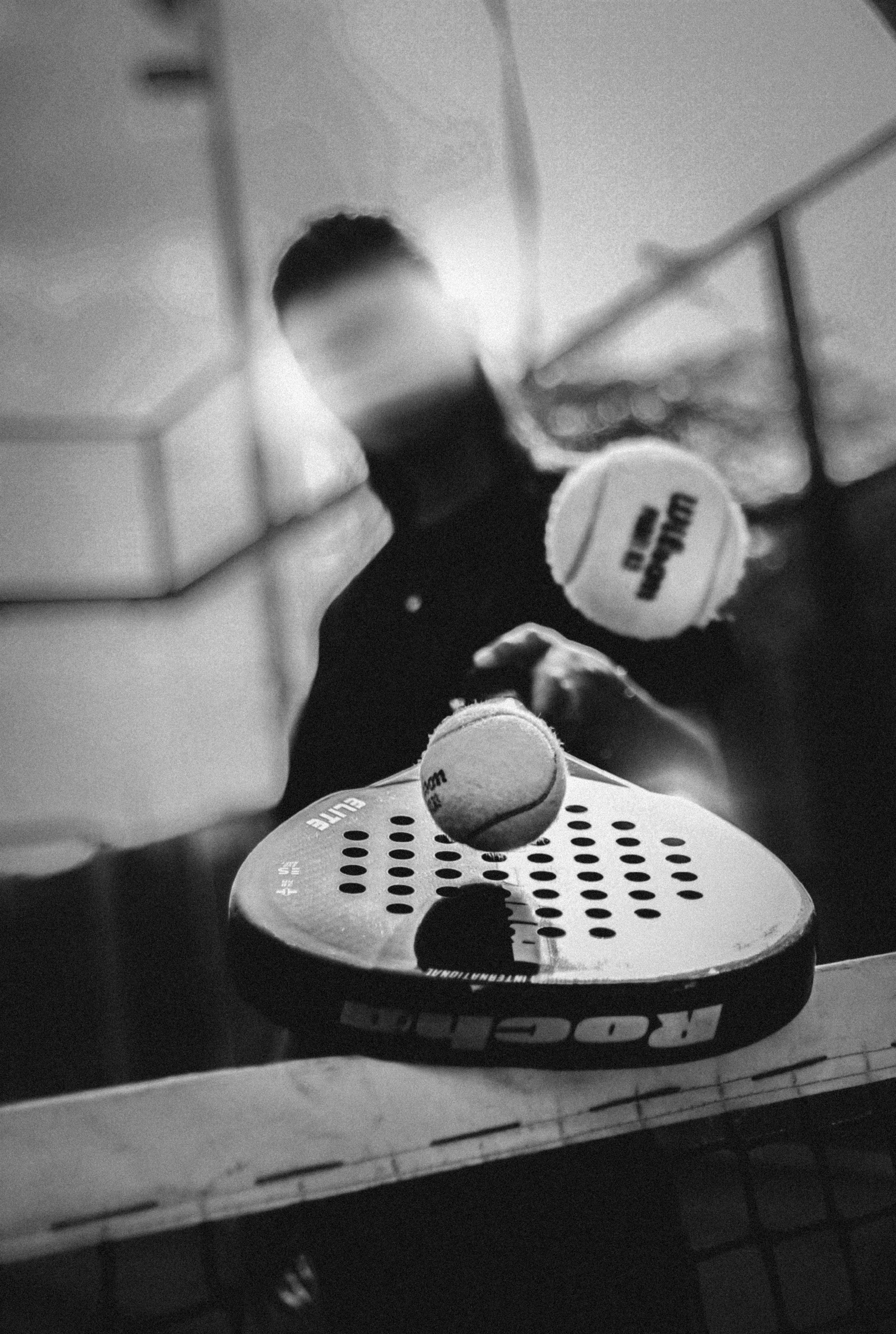 A blurred black and white image captures a person poised to strike a tennis ball with a paddle, emphasizing motion and focus in a padel court setting.