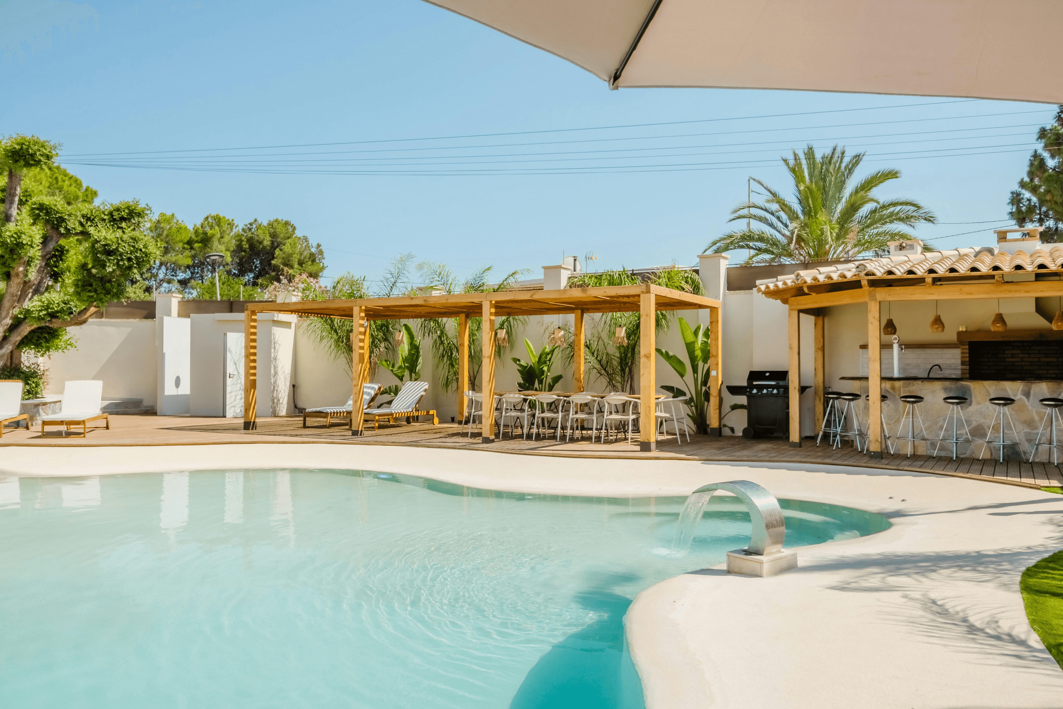 Outdoor pool area with pergola, lounge chairs, palm trees, and turquoise pool on a sandy patio.
