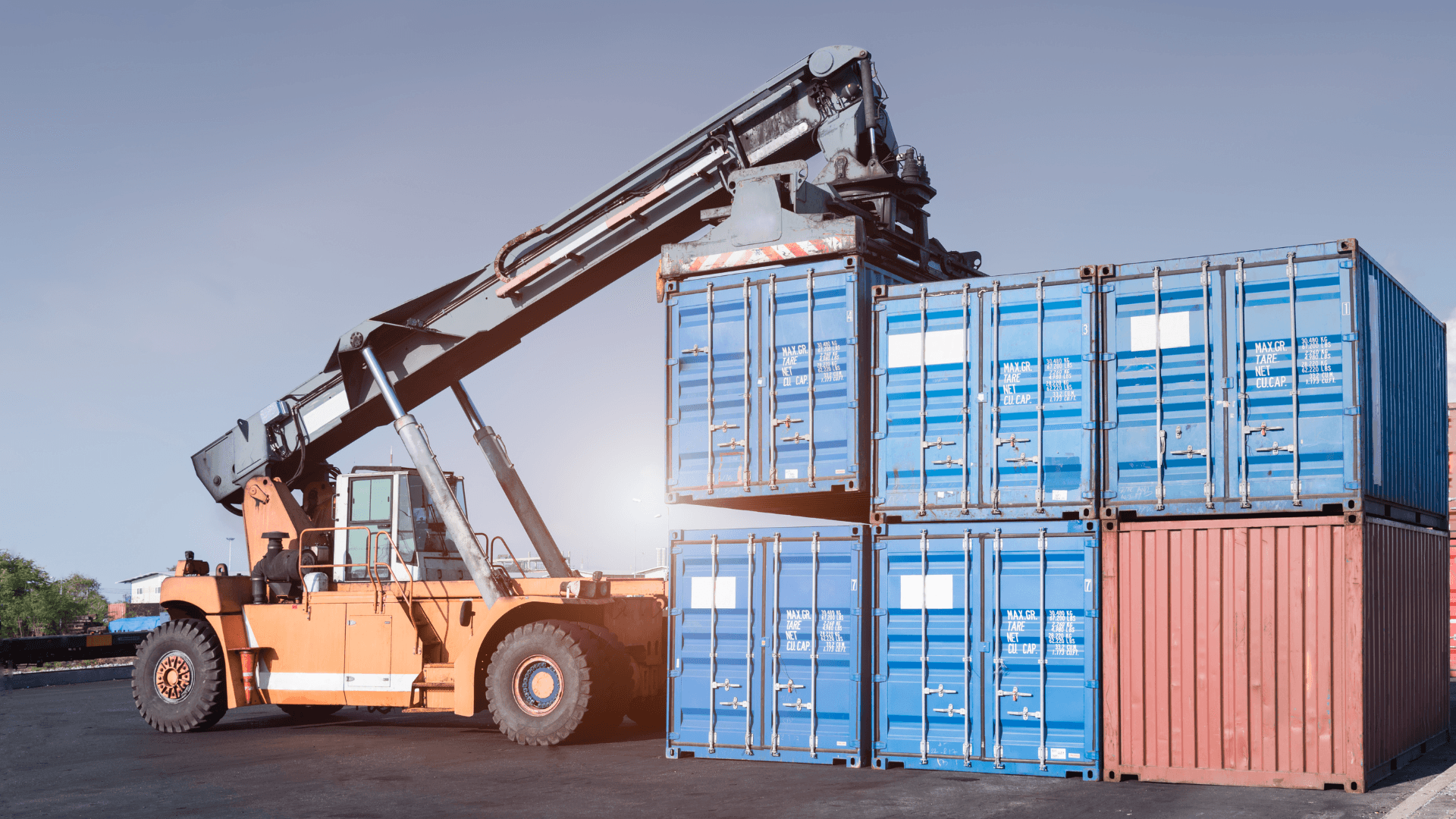 Reach stacker machine lifting blue shipping containers in a busy container yard.