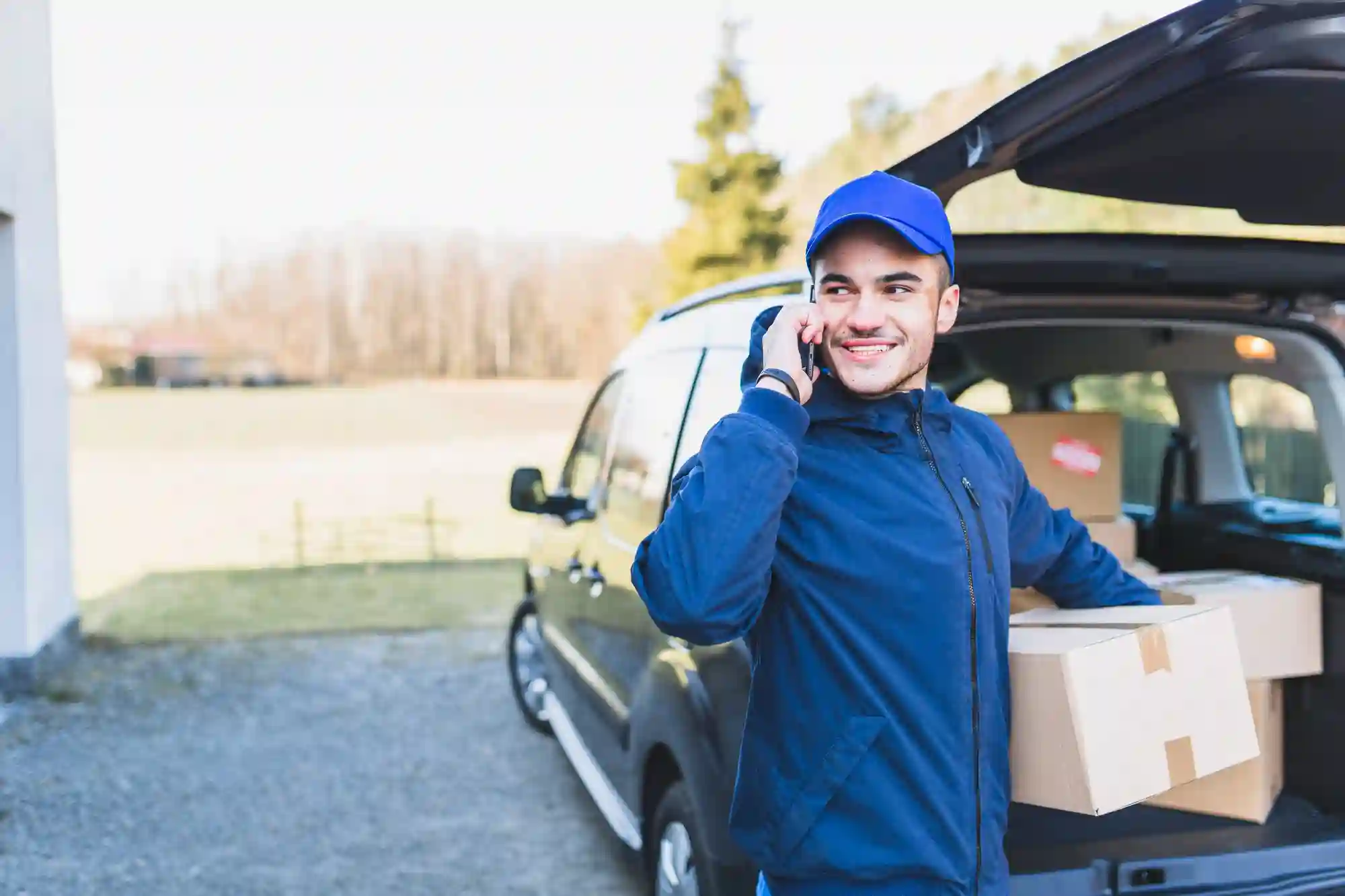 A smiling delivery driver talking on the phone while unloading cardboard packages from the trunk of a car.
