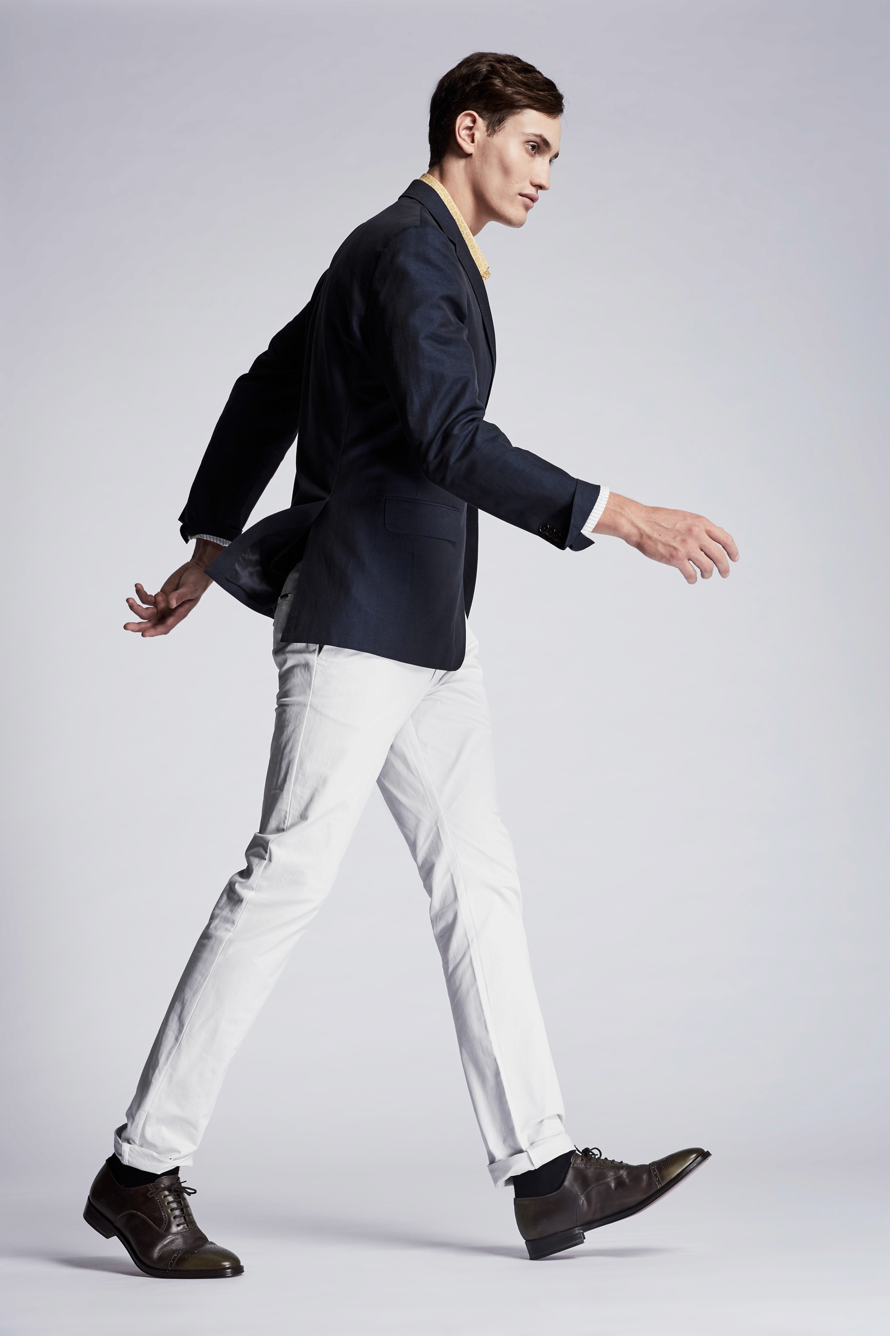 A side view of a male model striding, with swinging arms, across a minimal white background wearing a navy blazer and white pants with brown shoes.