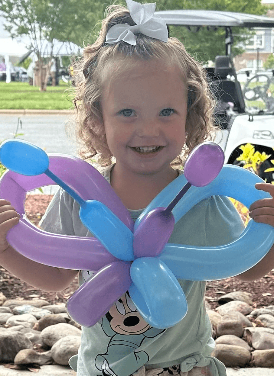 Smiling young girl holding a colorful balloon butterfly, standing outdoors with a cheerful background of trees and a golf cart