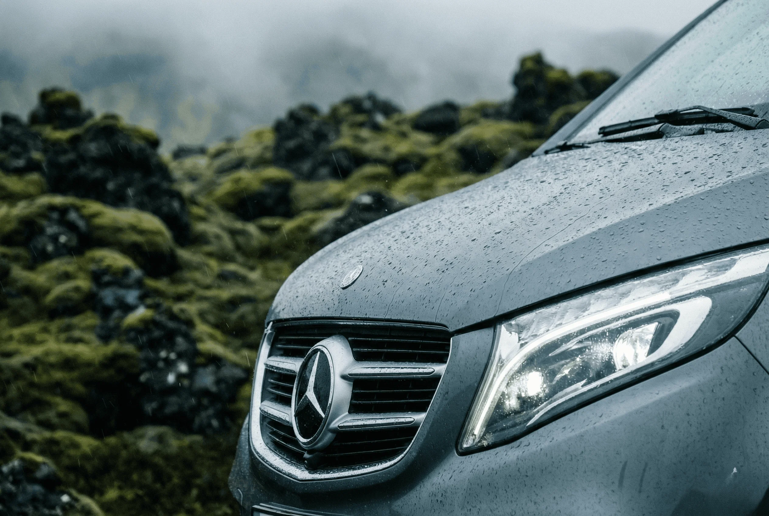 Close-up detail of the headlight and grille of a silver Mercedes van with water droplets, set against a mossy background.