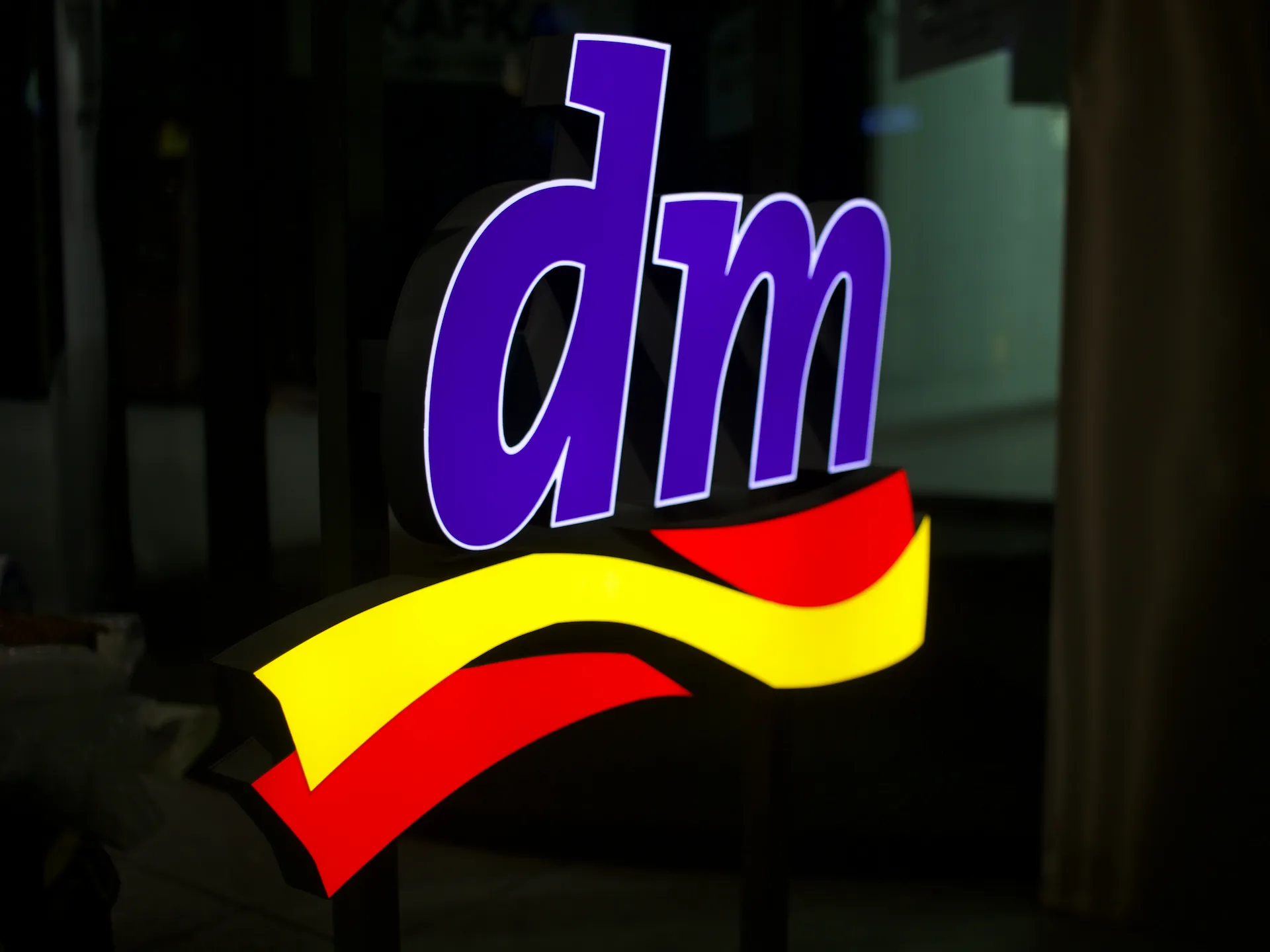 Barevné světelné logo dm drogerie – fialový nápis se žlutočervenou vlnou z akrylátu, noční záběr