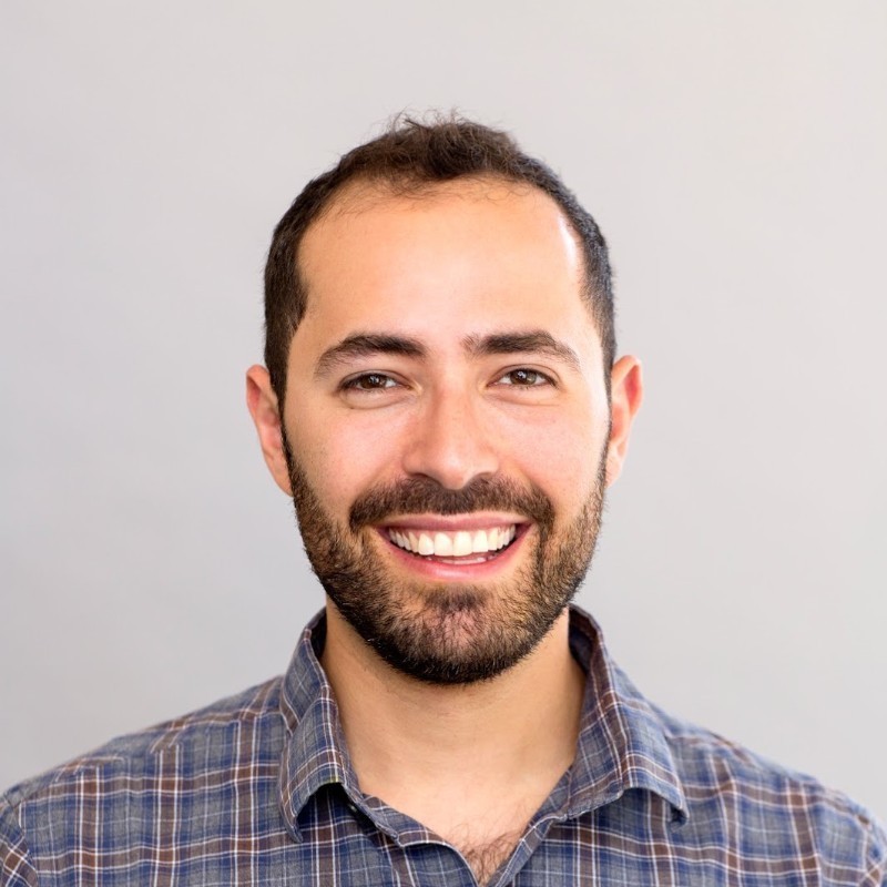 Smiling man with dark hair and a beard, wearing a plaid shirt.