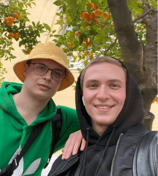 Pavlo and Maxym are smiling for a selfie under an orange tree, one wearing a yellow bucket hat and green hoodie, the other in a black leather jacket and hoodie.