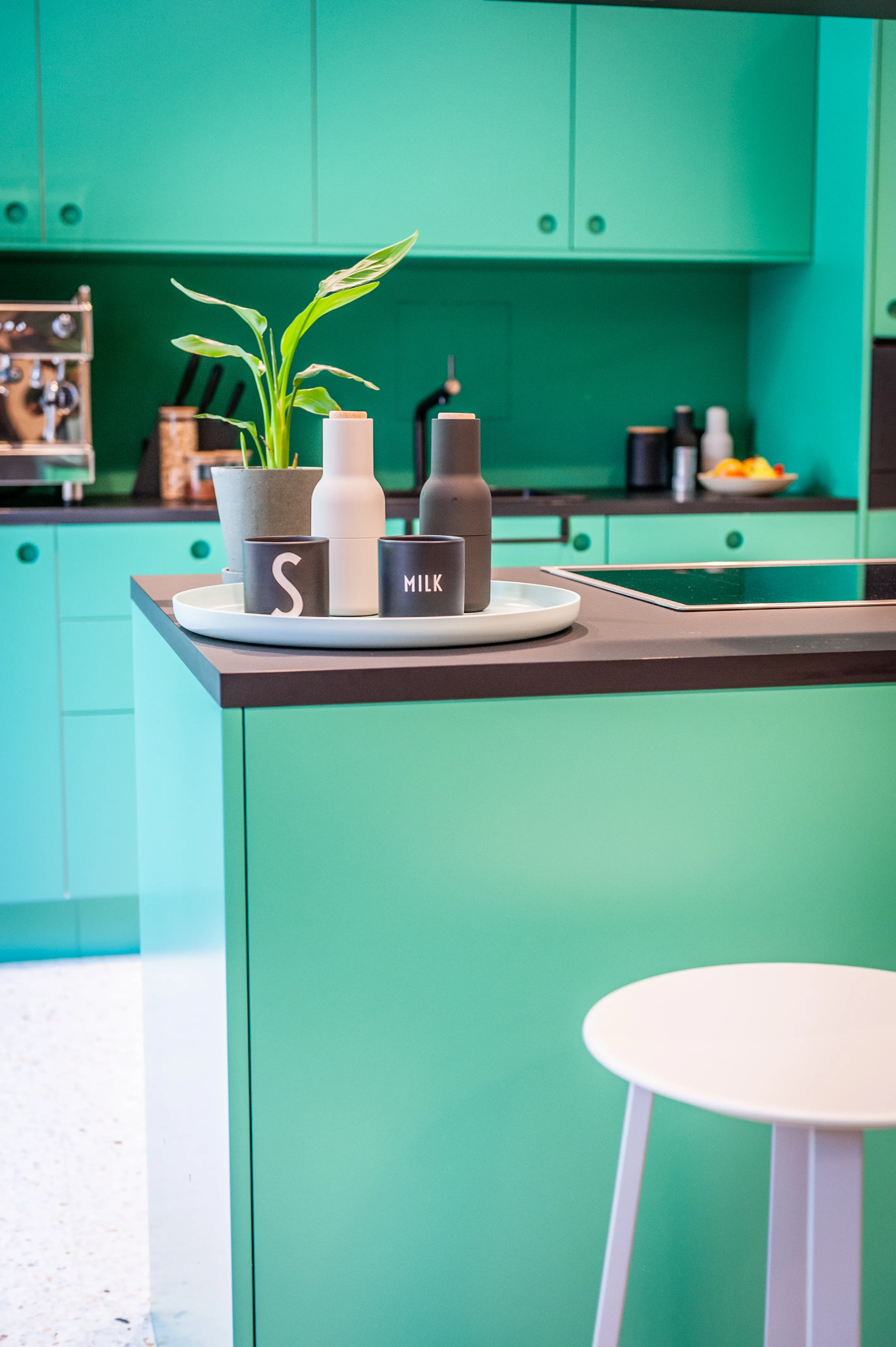 Detail of the mint-green kitchen island at EHEALTH-TEC with a dark countertop, a tray with designer jars and a milk carafe, a potted plant, espresso machine and a pale pink bar stool.