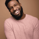 Headshot of a smiling Black man with a beard, wearing a pink sweater against a beige background.