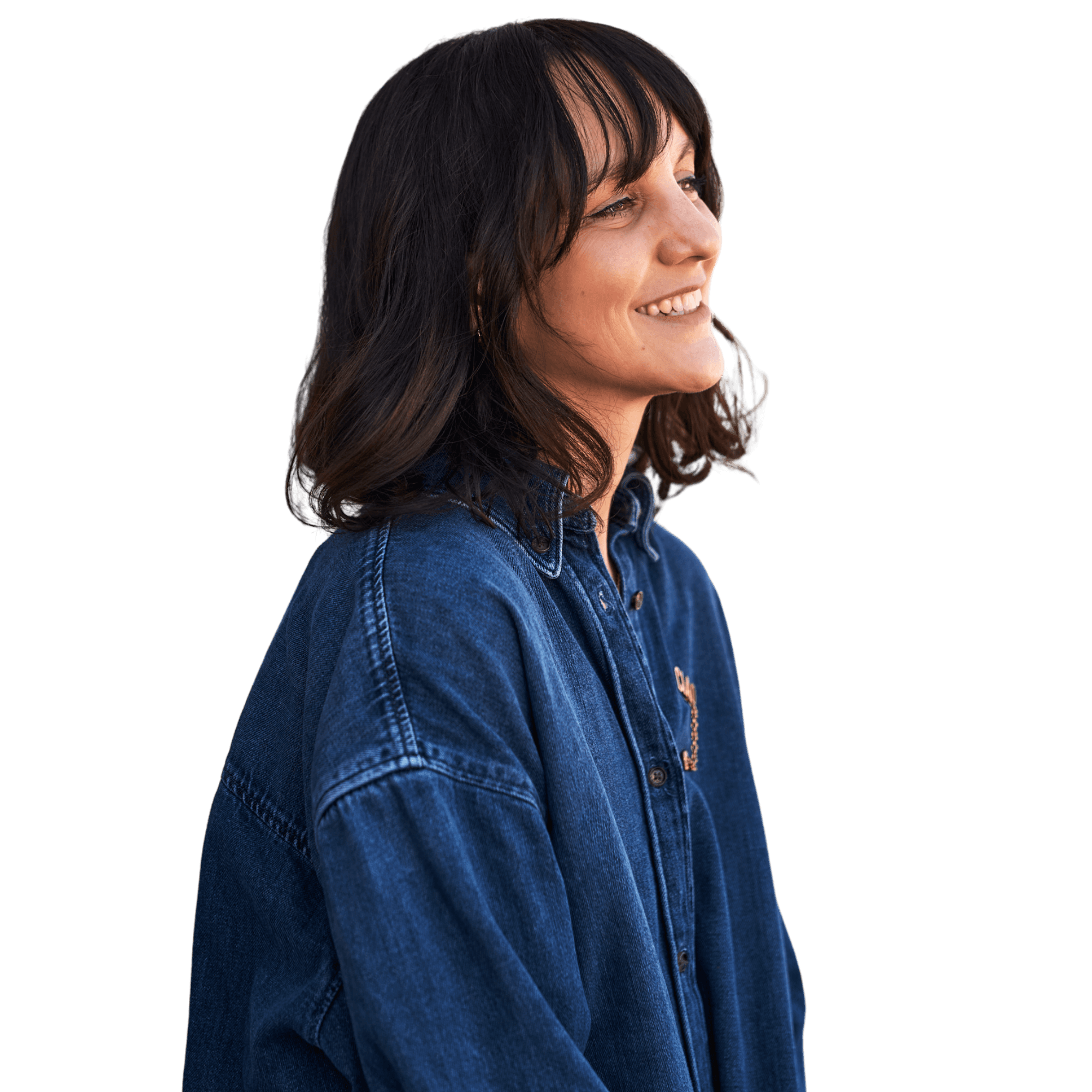 A young person with curly hair smiles at the camera while sitting in front of a laptop against a dark background.