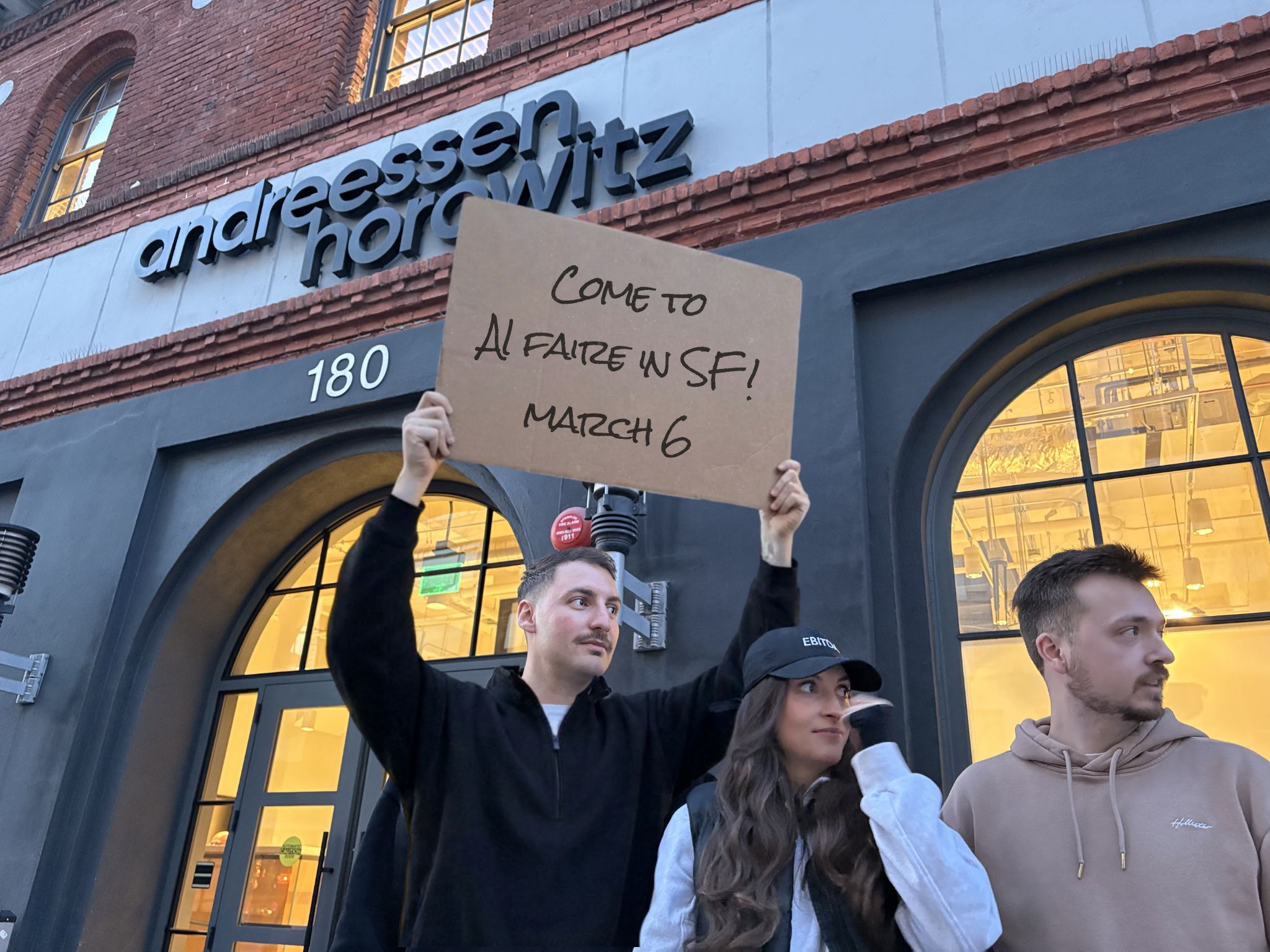 Co-Founder and CEO of Pluvo, Alexandre Labrèche, holds a sign reading “Come to AI Faire in SF! March 6” to promote the AI demo day. Co-founders Andrew Ingram and Vanessa Galarneau are next to him in front of the a16z building.