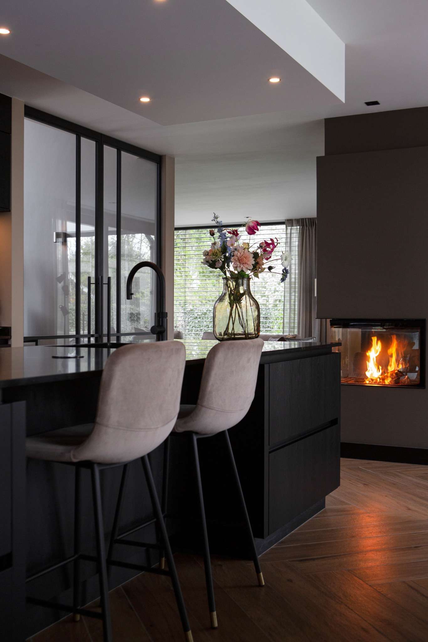 Keuken met zwart kookeiland, beige barstoelen en een glazen vaas met bloemen.