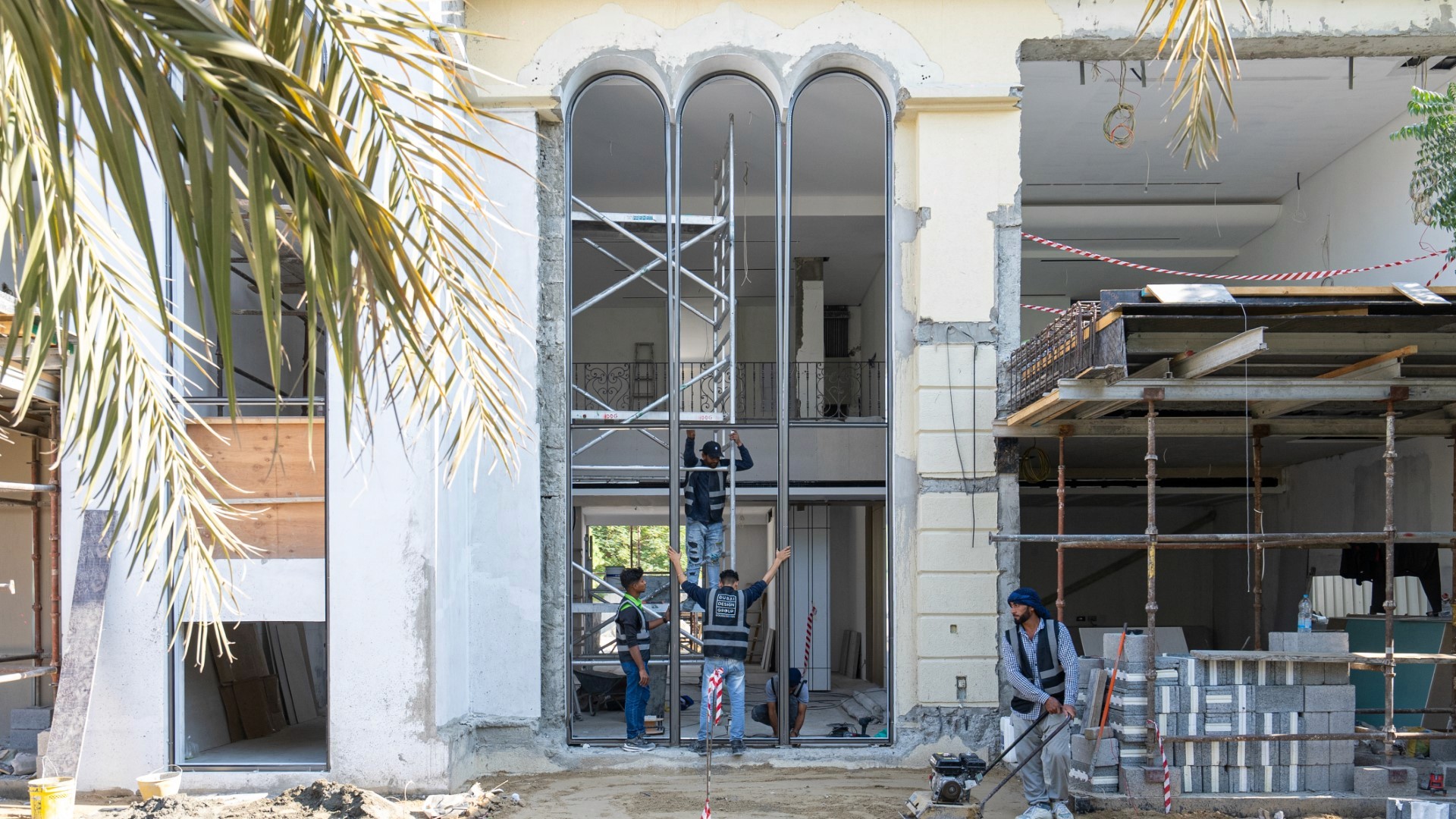 A villa in Jumeirah Islands under renovation, with scaffolding erected in front and construction workers actively engaged in various tasks; nearby, palm fronds add a touch of greenery to the scene.