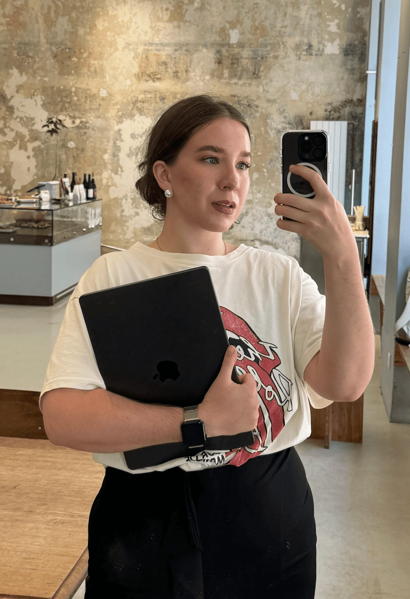 Digital strategist and co-founder of a creative agency, holding a laptop and taking a mirror selfie in a stylish café with rustic walls. Her calm, confident presence reflects the agency’s personalized, well-structured approach to social media and brand strategy.