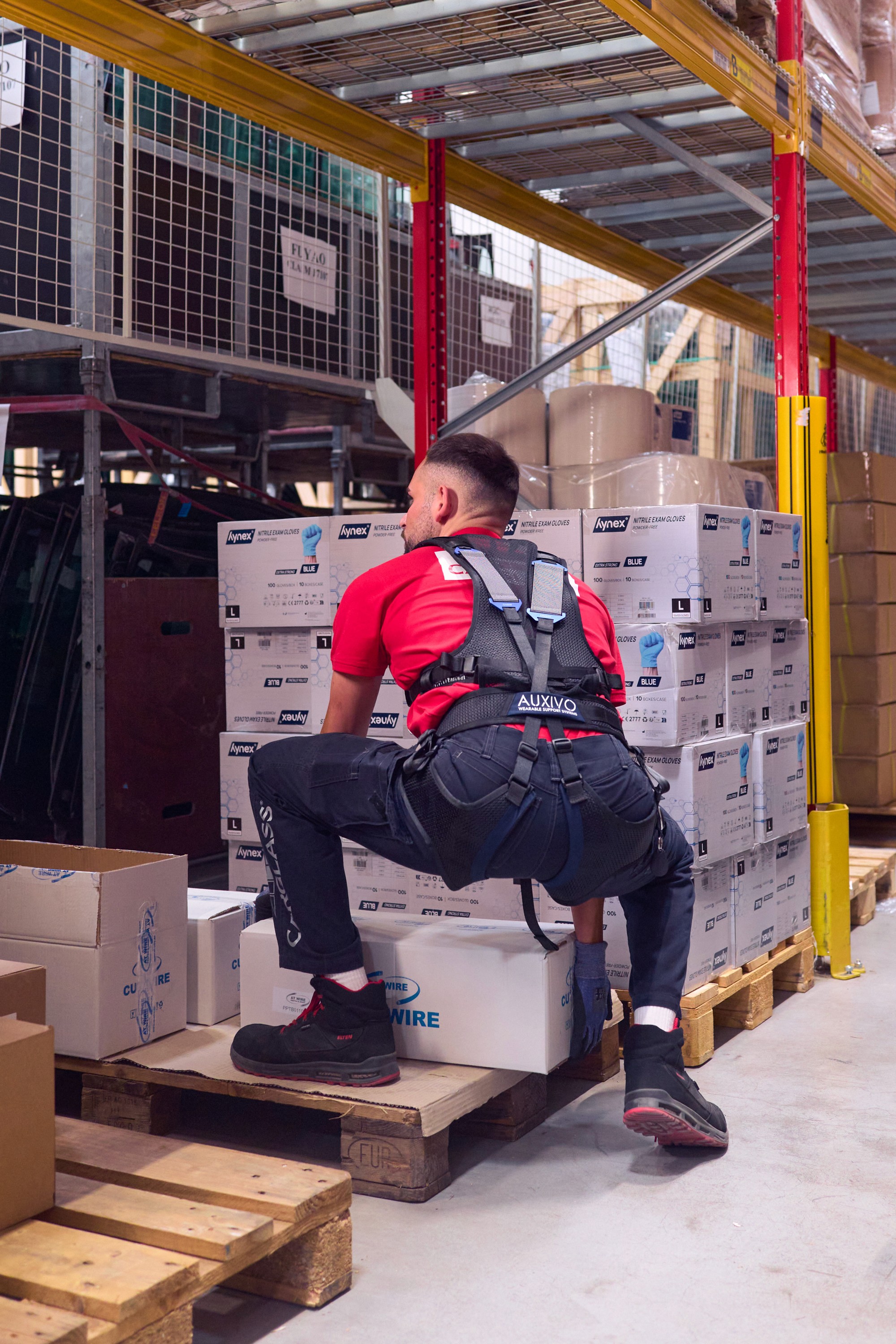 Travailleur d’entrepôt soulevant une boîte au sol en portant l’exosquelette LiftSuit d’Auxivo, conçu pour réduire la charge sur le bas du dos lors des tâches de manutention.