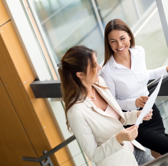 A young person with curly hair smiles at the camera while sitting in front of a laptop against a dark background.