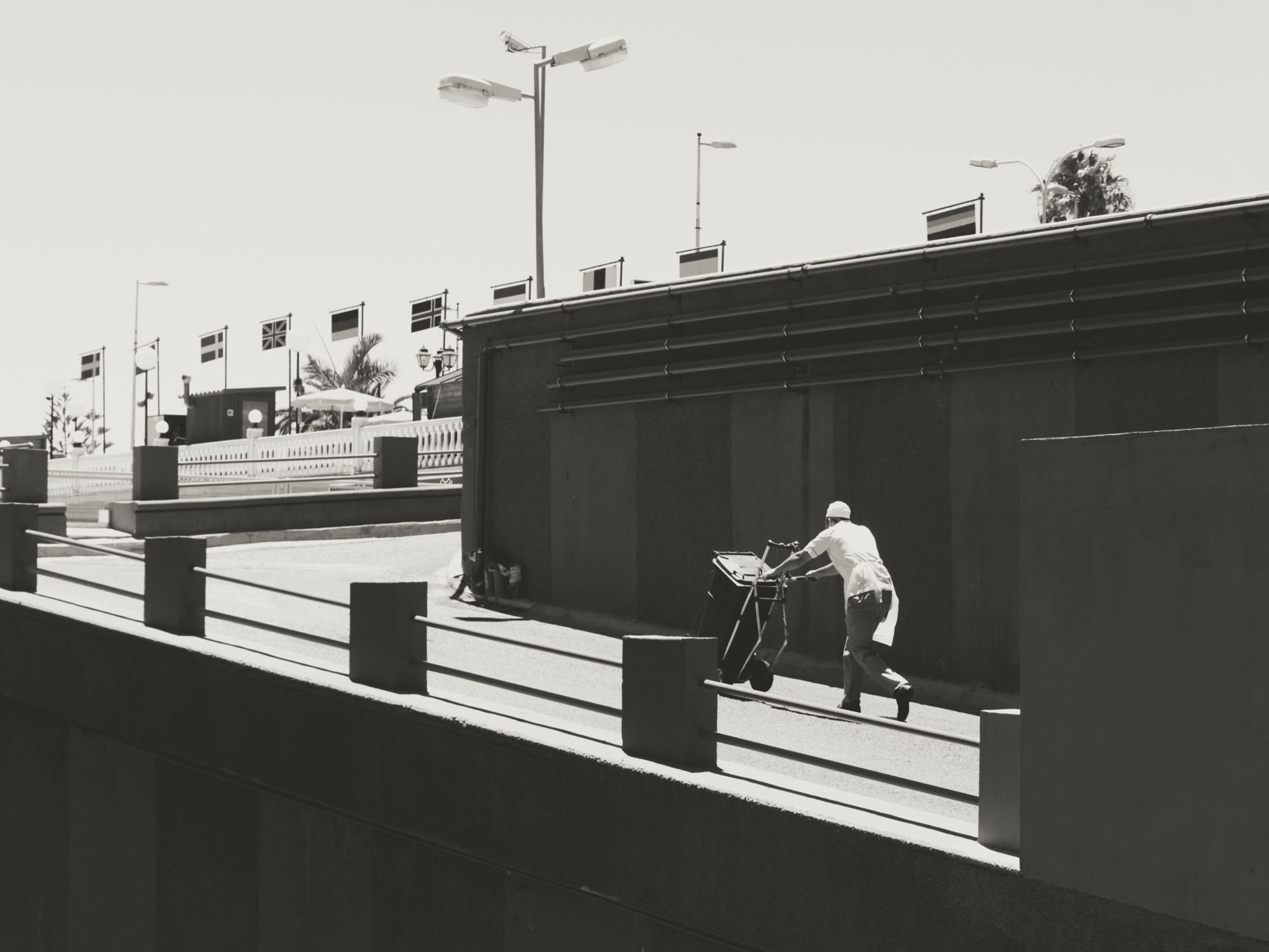 High-contrast urban scene showing a lone figure pushing a cart uphill on a ramp, emphasizing geometric lines, shadows, and a distant row of flags and palm trees.