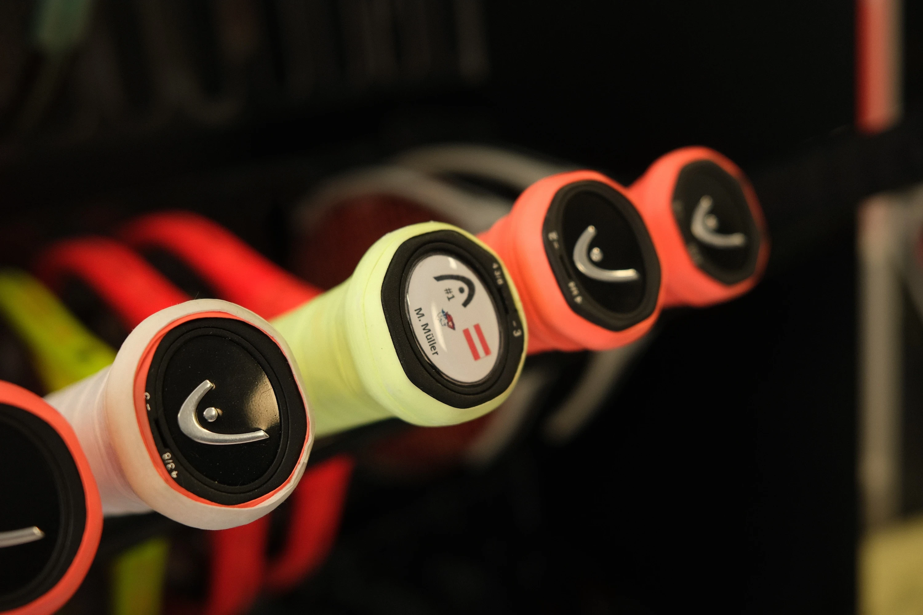 Mehrere Tennisschläger mit Griffbändern in verschiedenen Farben, darunter Neon-Gelb und Koralle, stehen in einer Halterung. Die Schlägerenden zeigen das Logo der Marke HEAD, und auf einem Griff ist eine kleine österreichische Flagge zu sehen. Der Hintergrund ist unscharf, während die Griffe im Vordergrund im Fokus stehen.