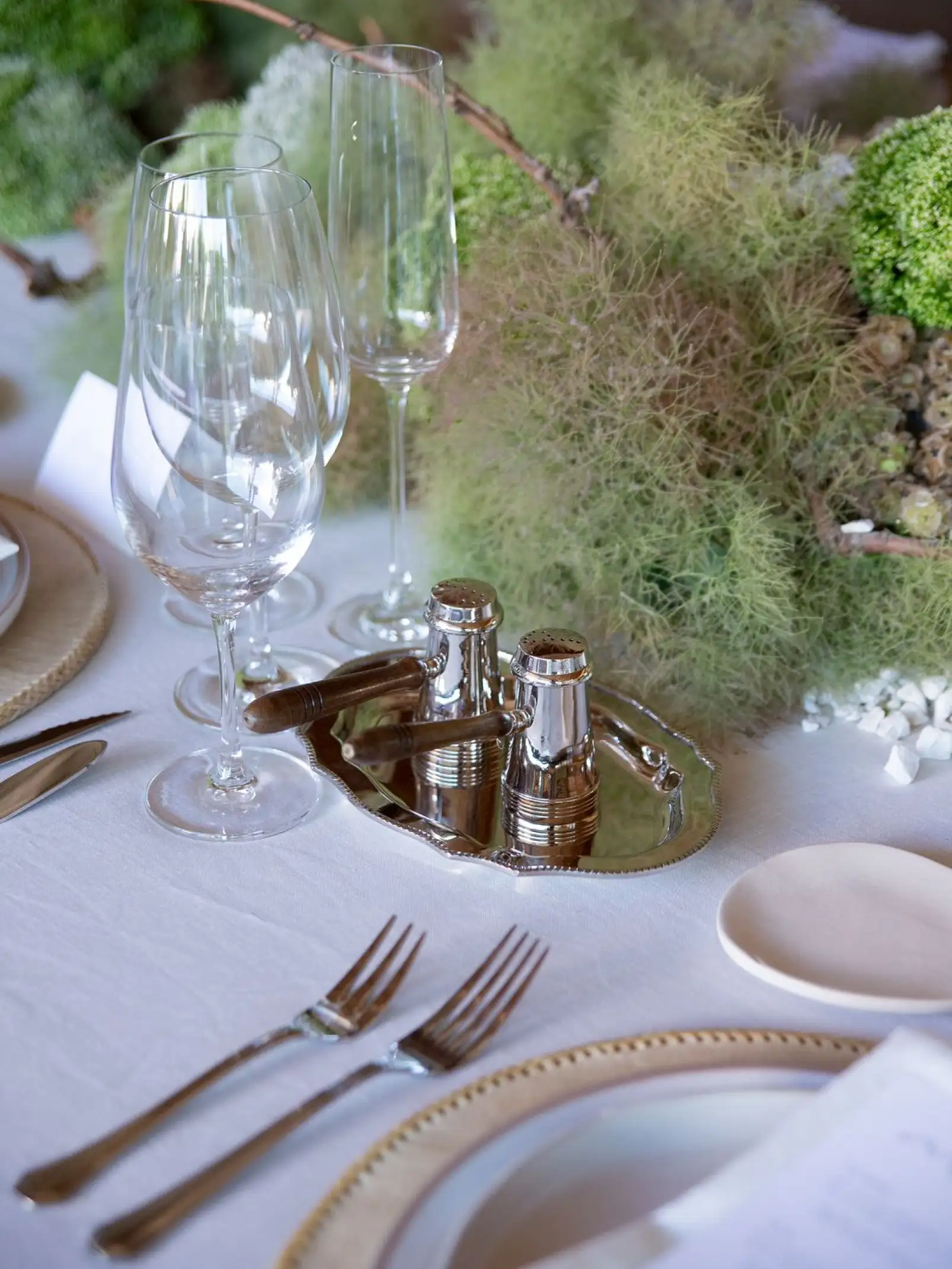 Lugar de mesa con platos blancos, servilleta y menú, rodeado de decoración vegetal minimalista en tonos verdes y neutros.