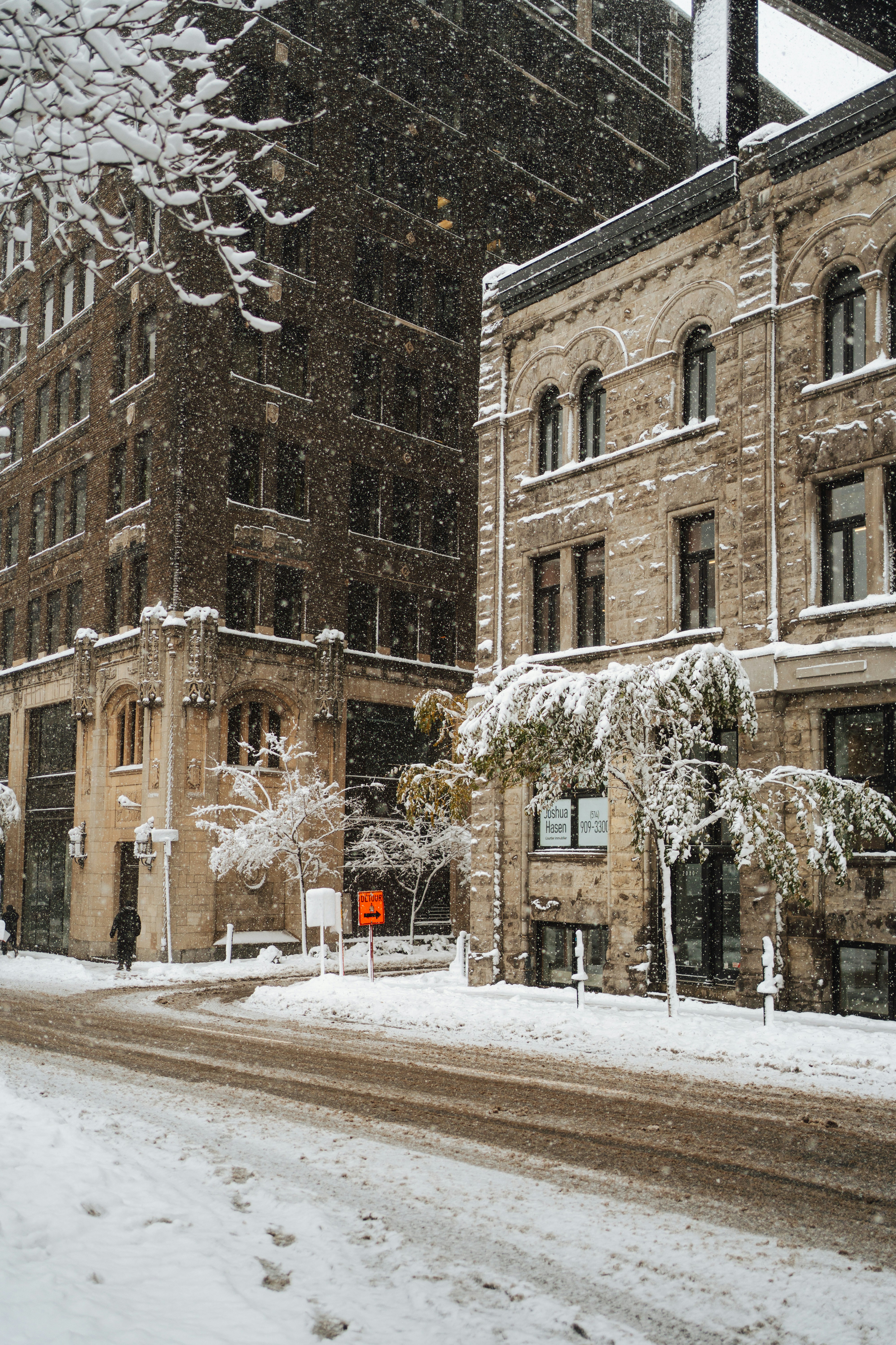 Snow falls on a city street with buildings.
