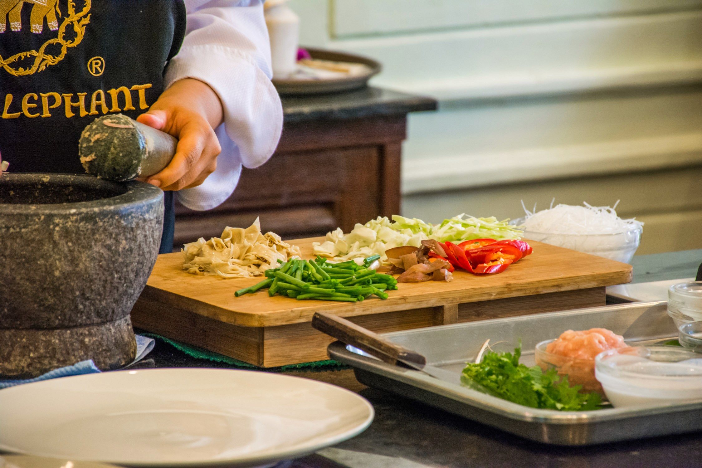 A chef's hands use a mortar and pestle to grind spices next to sliced vegetables on a wooden board in a kitchen.