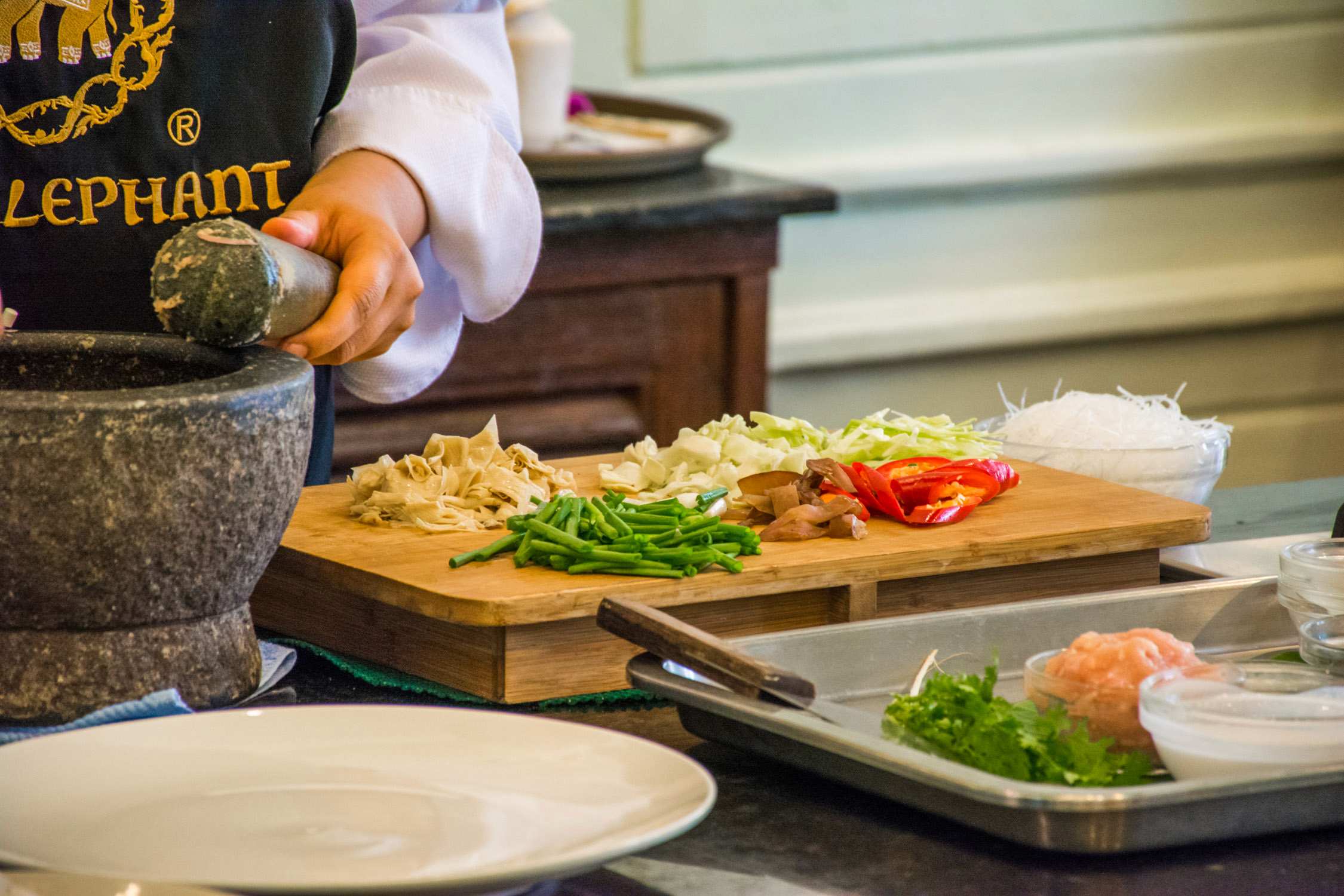 A chef's hands use a mortar and pestle to grind spices next to sliced vegetables on a wooden board in a kitchen.