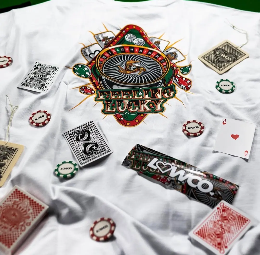 White t-shirt featuring Lowco Automotive Feeling Lucky graphic with casino-inspired illustration, playing cards, poker chips, and roulette wheel artwork.