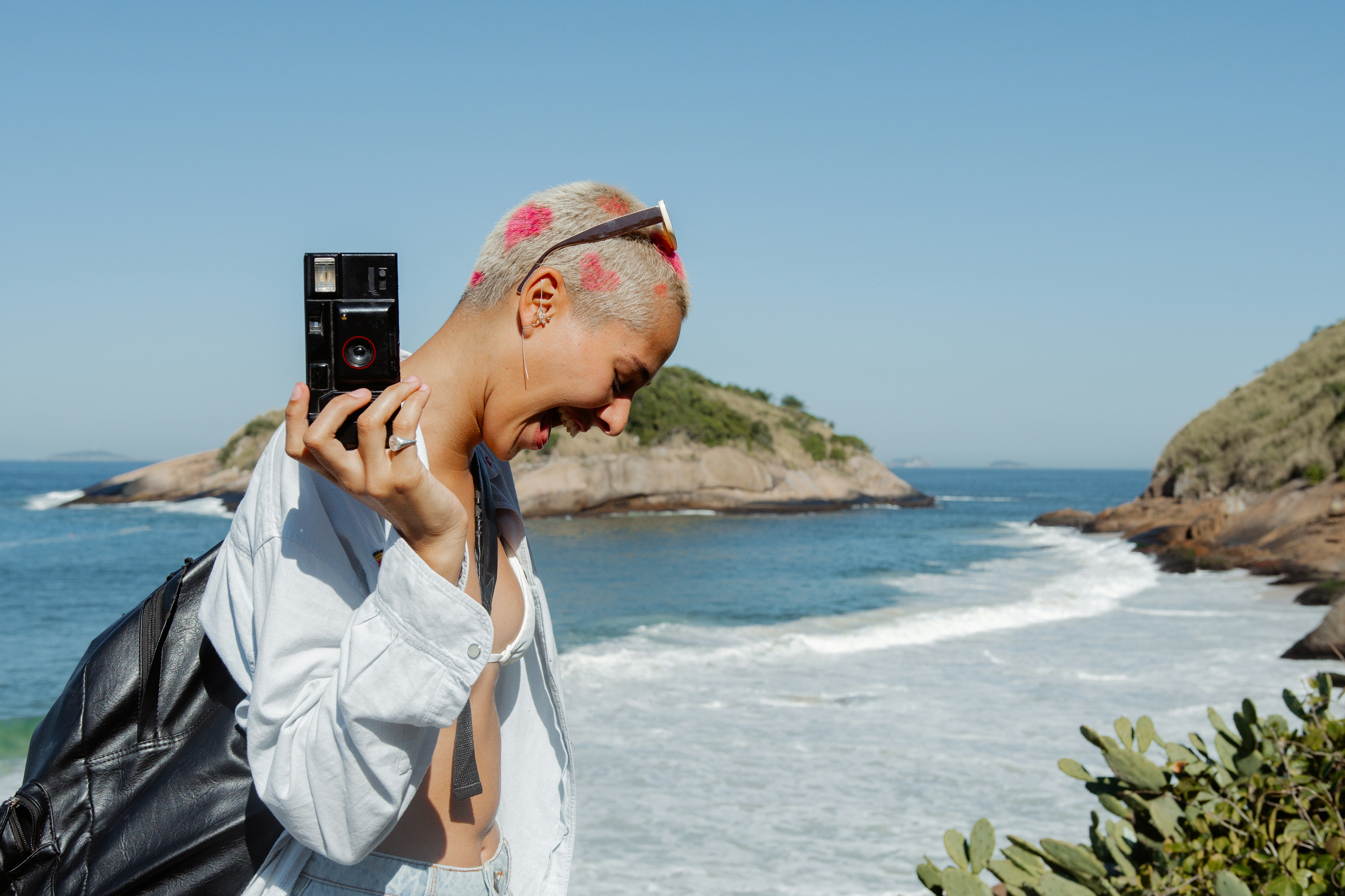A woman holds a camera, smiling with her tongue out, while standing on the beach with the water in the background.