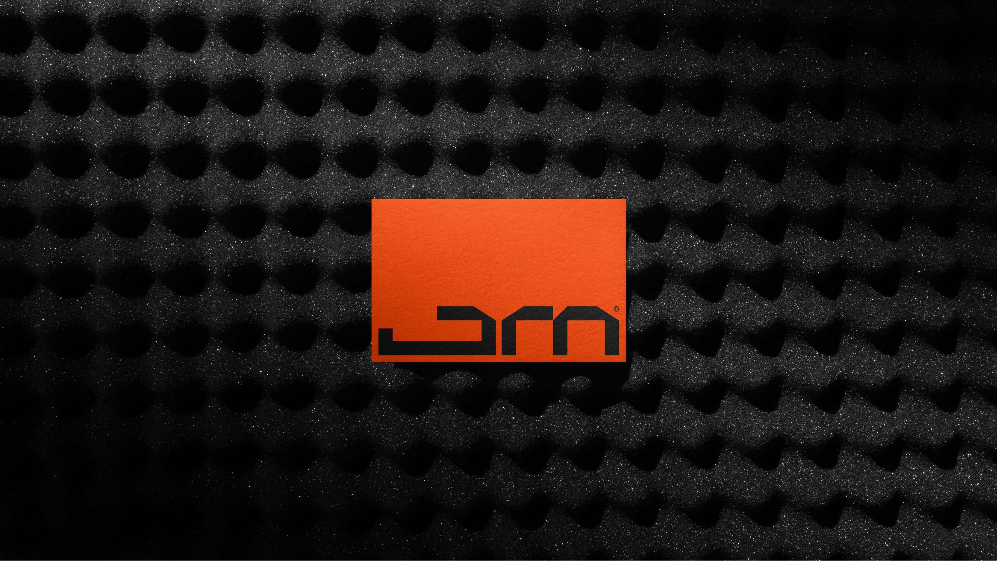 Person wearing black clothing carrying a black tote bag with a green globe-like geometric symbol, standing against a concrete wall in sunlight and shadow.