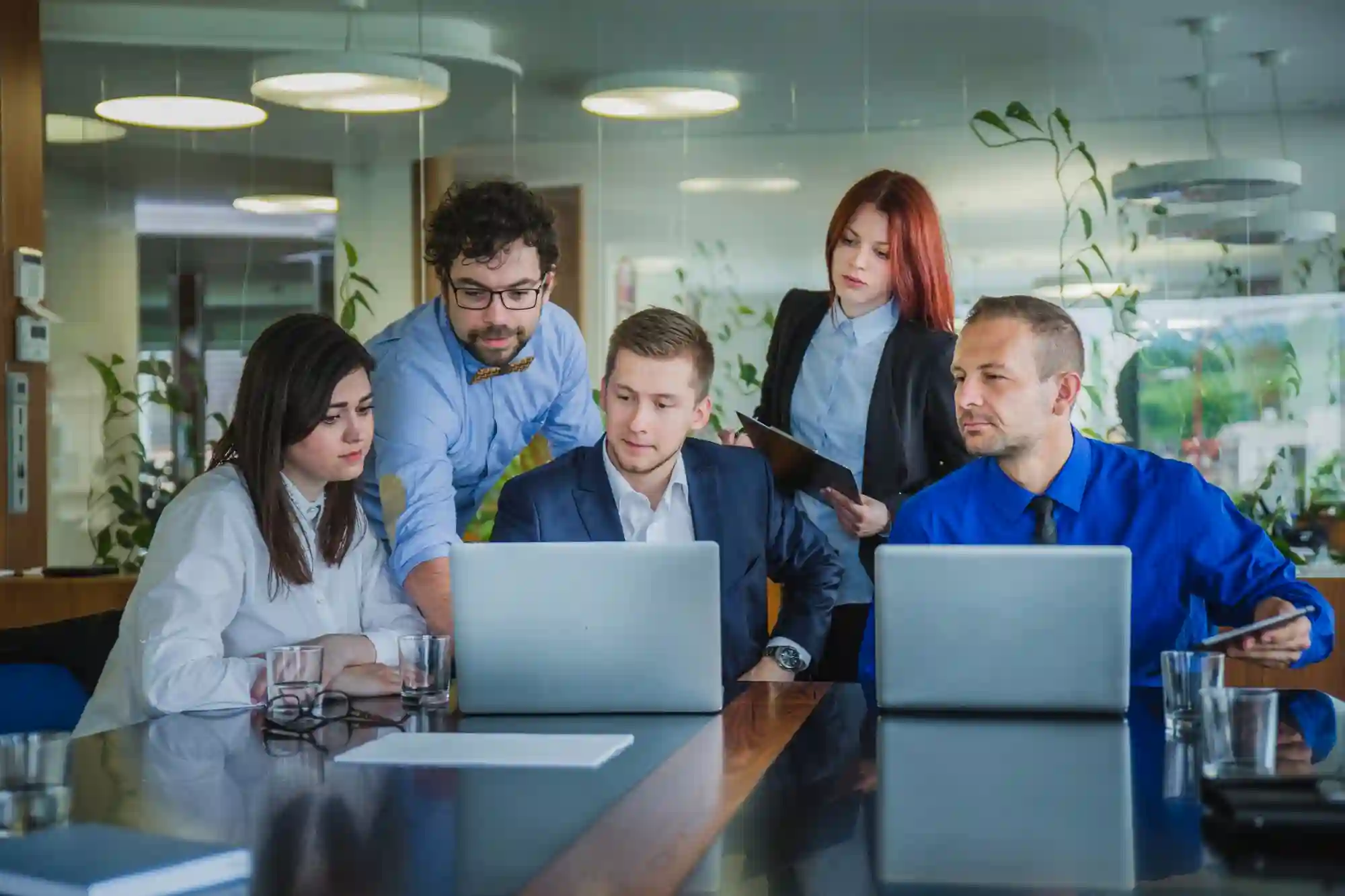 A diverse team of corporate professionals gathers around a laptop in a modern office to review project data.
