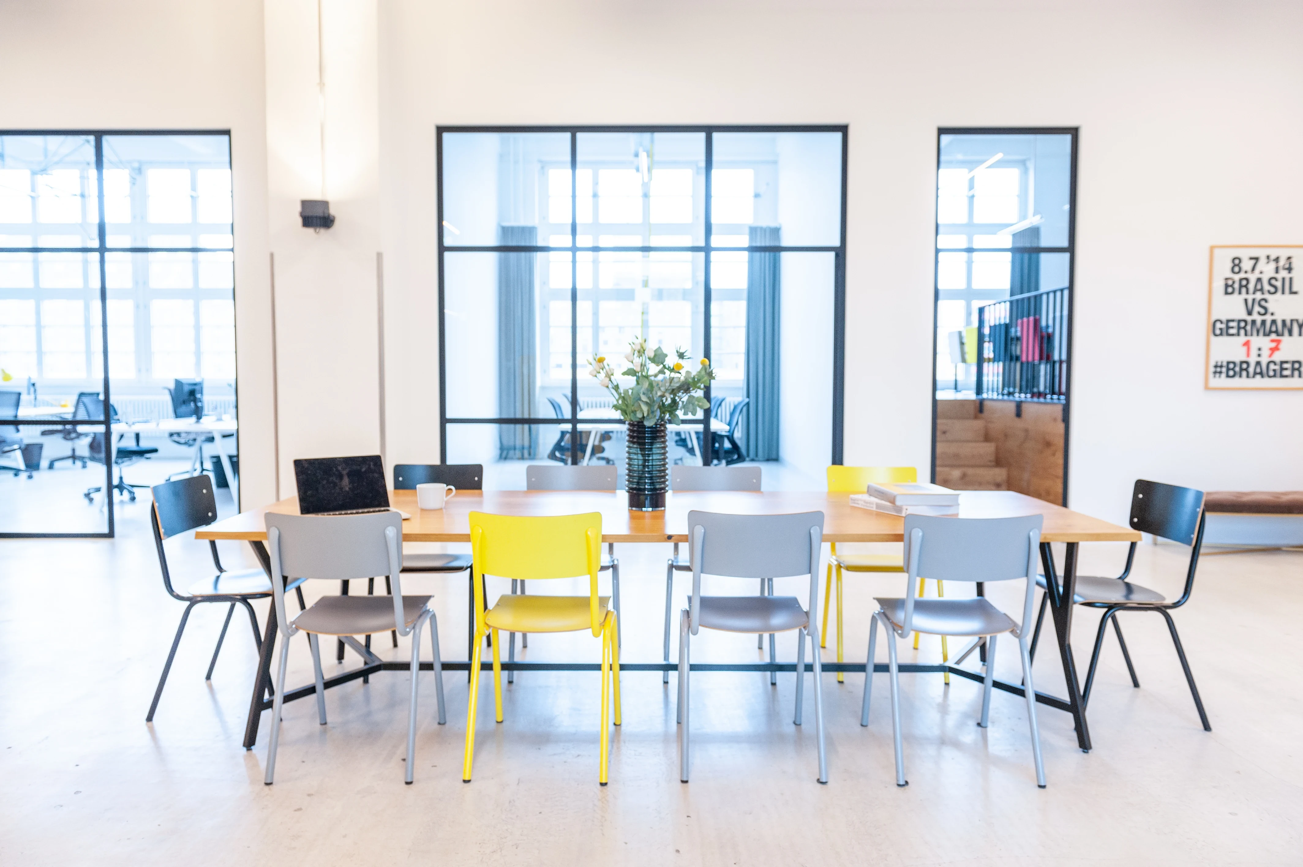 Canteen table at LA RED with a wooden top and mixed grey, black and yellow chairs, flowers in a vase, black-framed glass walls and a Brazil vs. Germany poster in the background.
