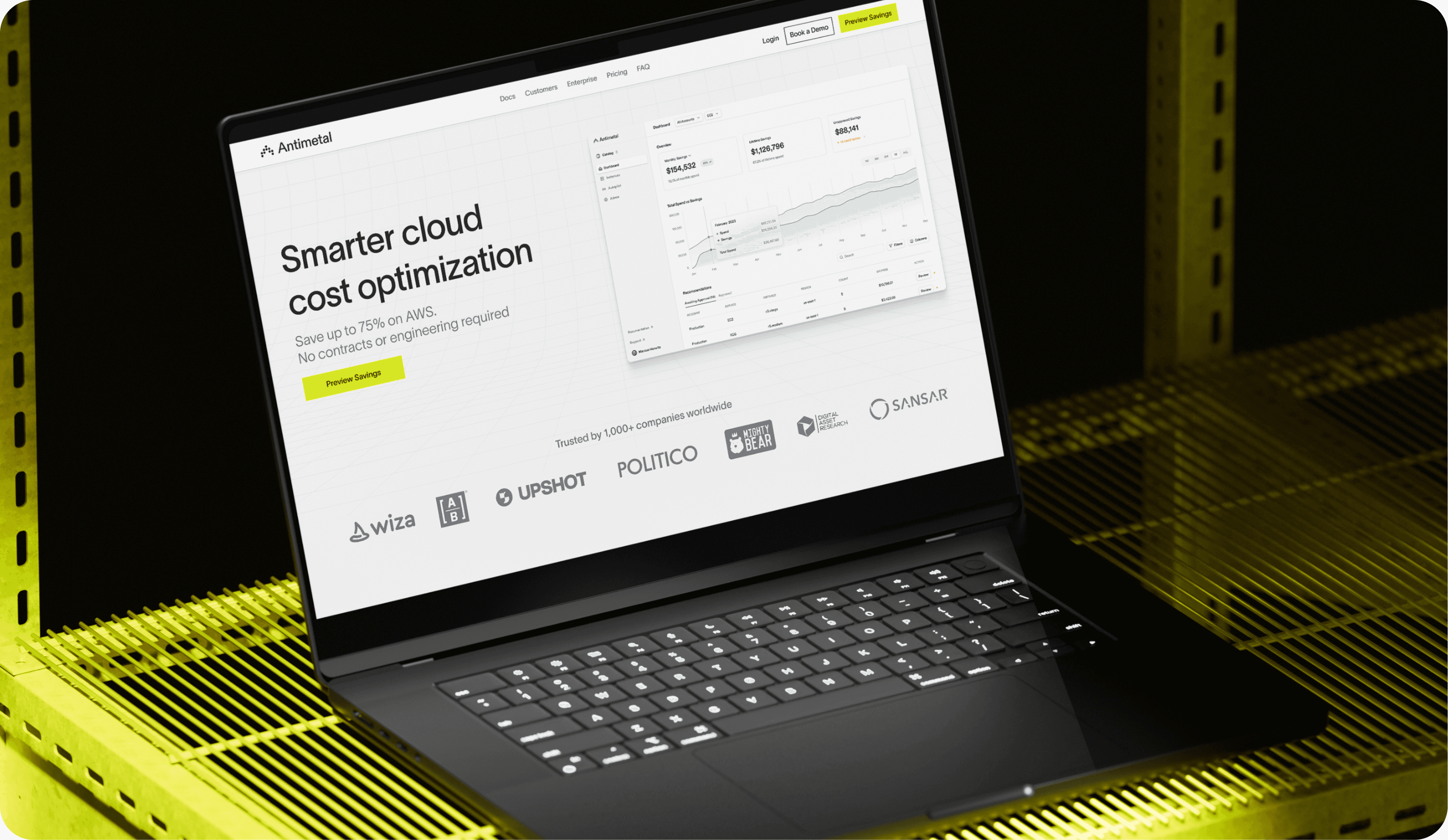 Black laptop displaying Antimetal's website on a yellow wireframe surface. The screen highlights 'Smarter cloud cost optimization' with the claim of saving up to 75% on AWS. Trusted by companies like Wiza, Upshot, Politico, and others, the website invites users to preview savings.