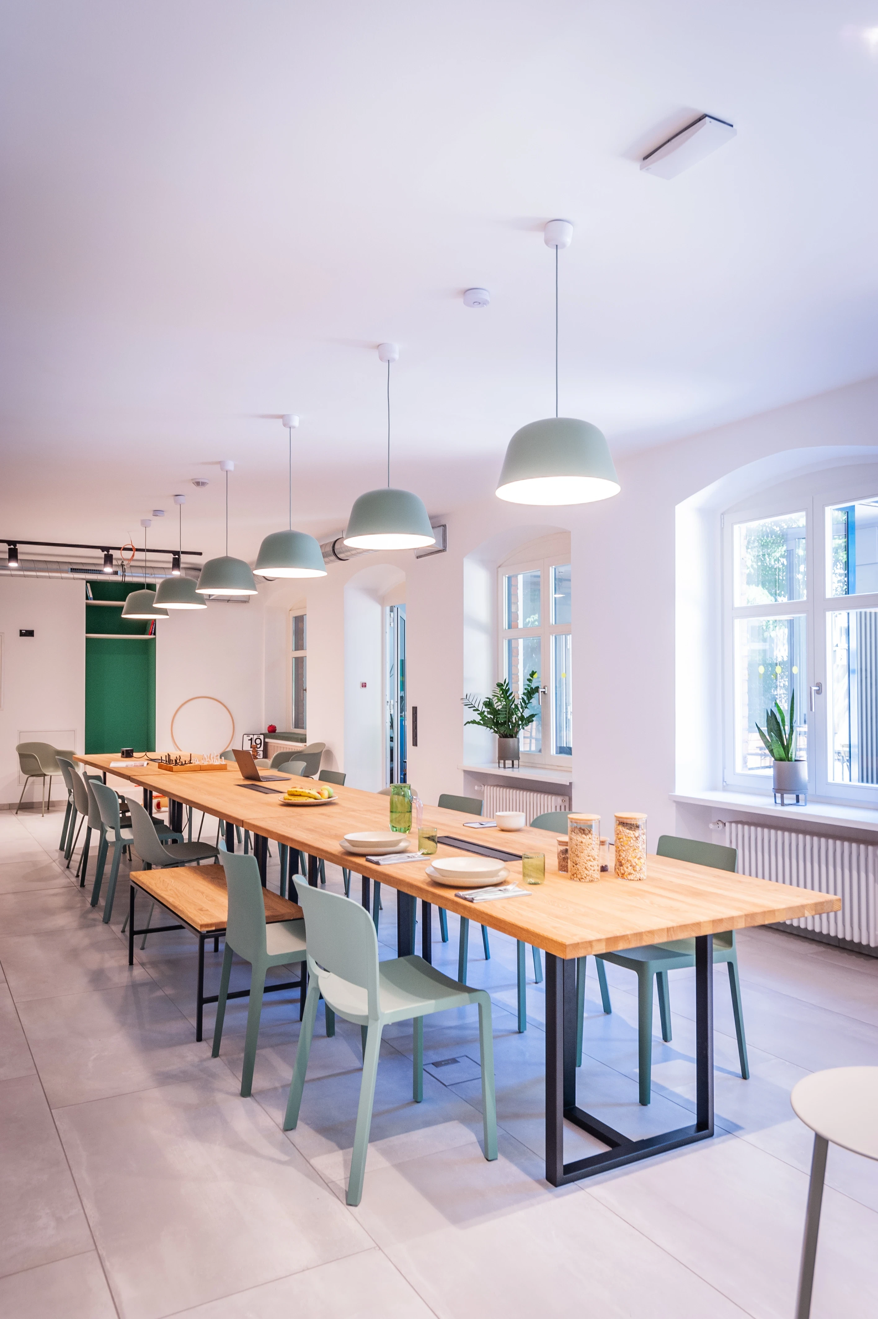 Long canteen table at EHEALTH-TEC made of butcher-block wood on black steel frames, with sage-green chairs and benches, matching green dome pendant lights and arched windows.