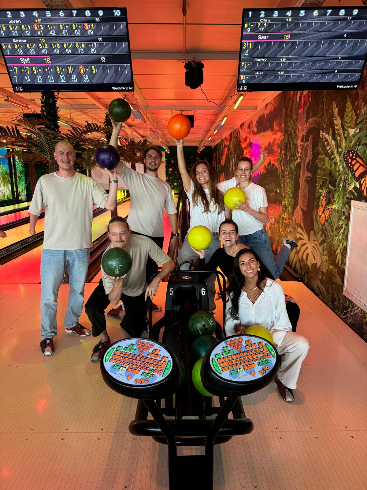 A group of friends smiles together in a bowling alley, with colorful lights and lanes in the background.