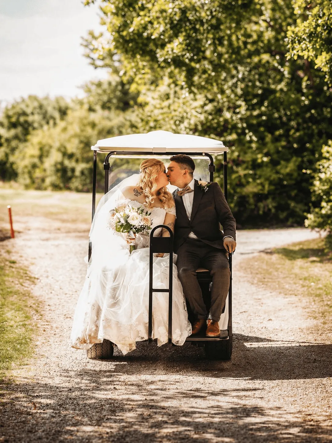 Newlyweds kiss in a golf cart on a sunny day. The bride wears a gown & veil, holding flowers.