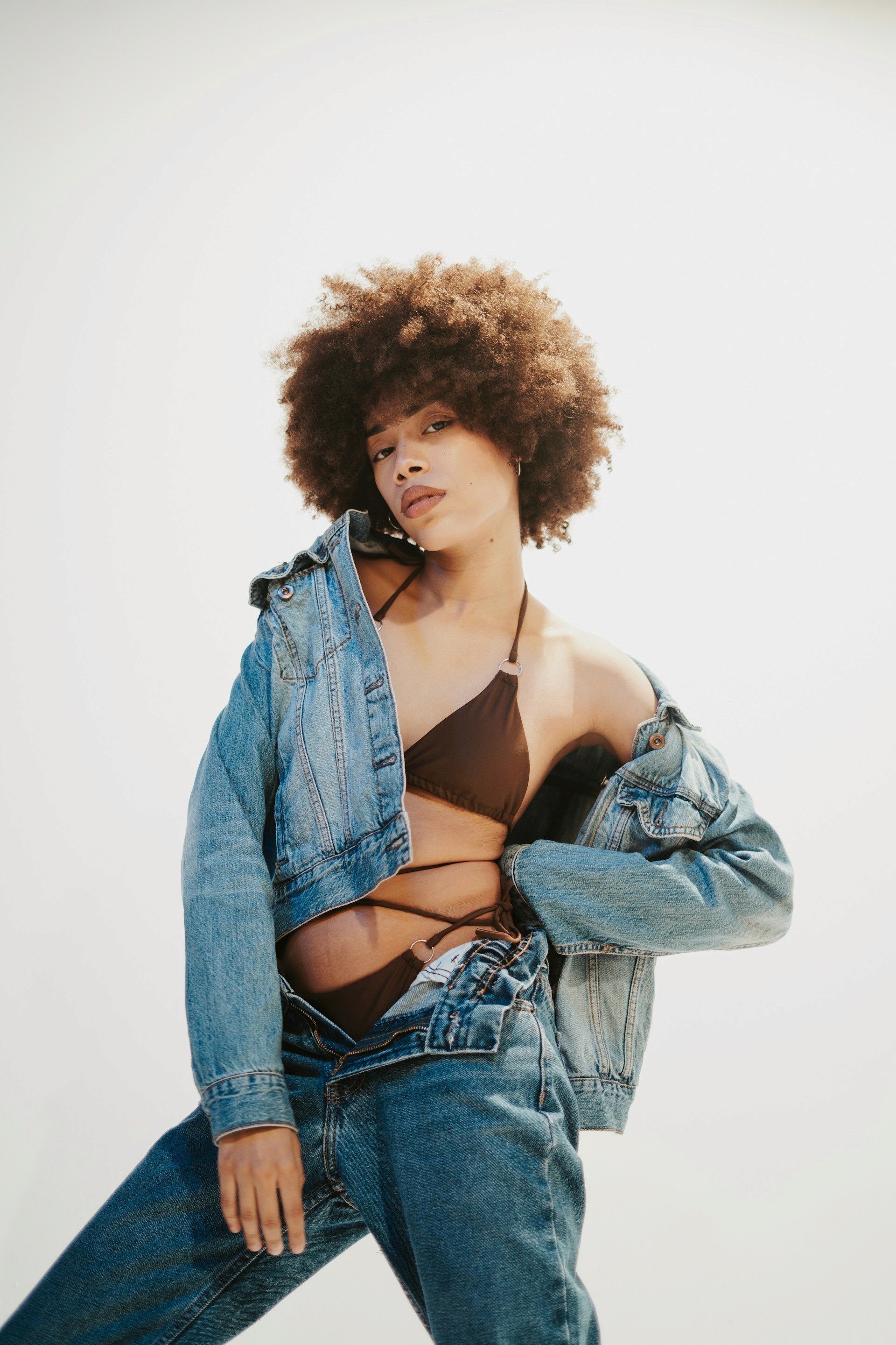 A woman with a large brown afro posing in a brown bikini top and an oversized denim jacket, with her matching denim jeans unbuttoned.
