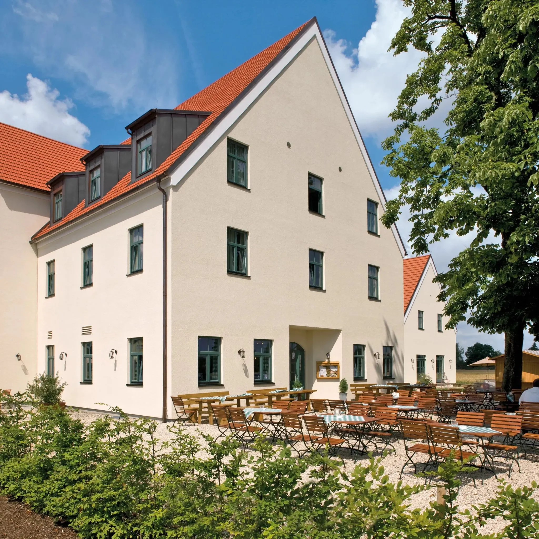 Außenansicht eines hellen Gebäudes mit roten Dachziegeln und vielen Tischen und Stühlen im Freien.