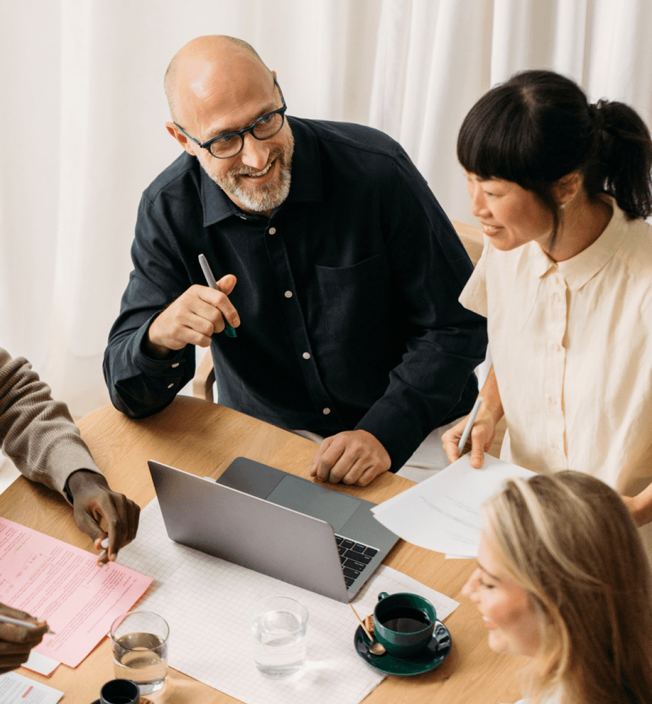 Team of diverse colleagues smiling and discussing around a laptop at a meeting table.