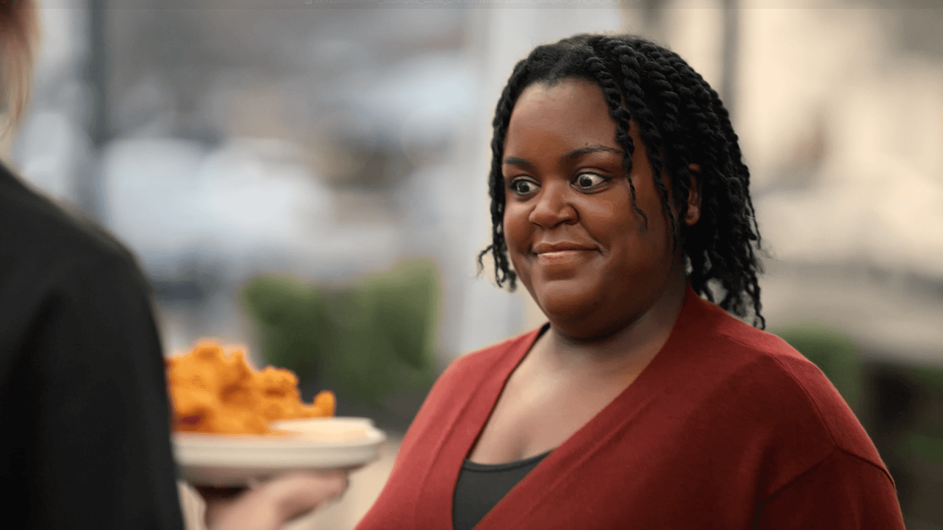 A smiling woman in a maroon sweater holds a plate of food, engaging with a person in the foreground.