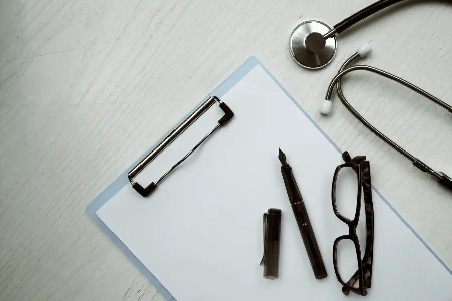 AI medical coding documentation represented by a blank clipboard, fountain pen, reading glasses, and stethoscope arranged on a white wooden desk.