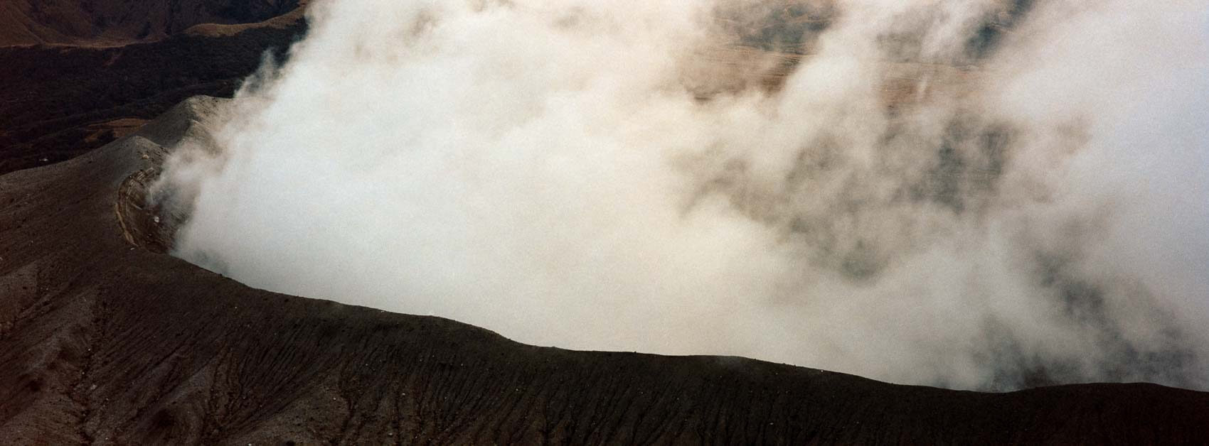 Japan hiking at Mount Aso: the crater spitting fumes, dramatic unworldly rough volcanic landscape, with majority clouds and showing some people through the clouds, captured on film in Kyushu's otherworldly terrain.