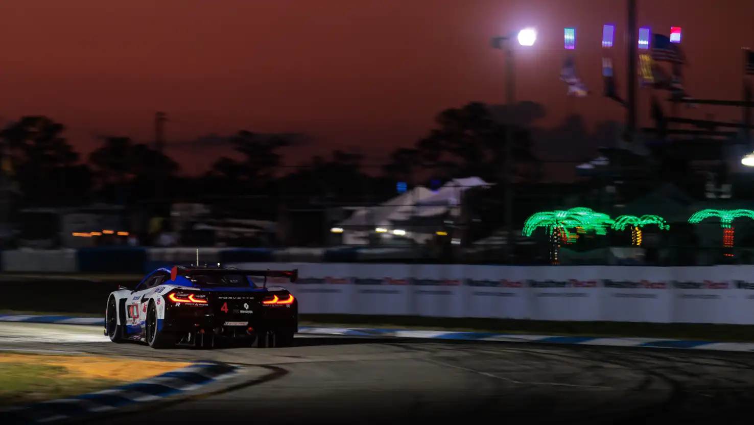 A Corvette race car speeds through a corner at Sebring at dusk, with glowing neon palm trees and dark silhouettes of fans and tents in the background.