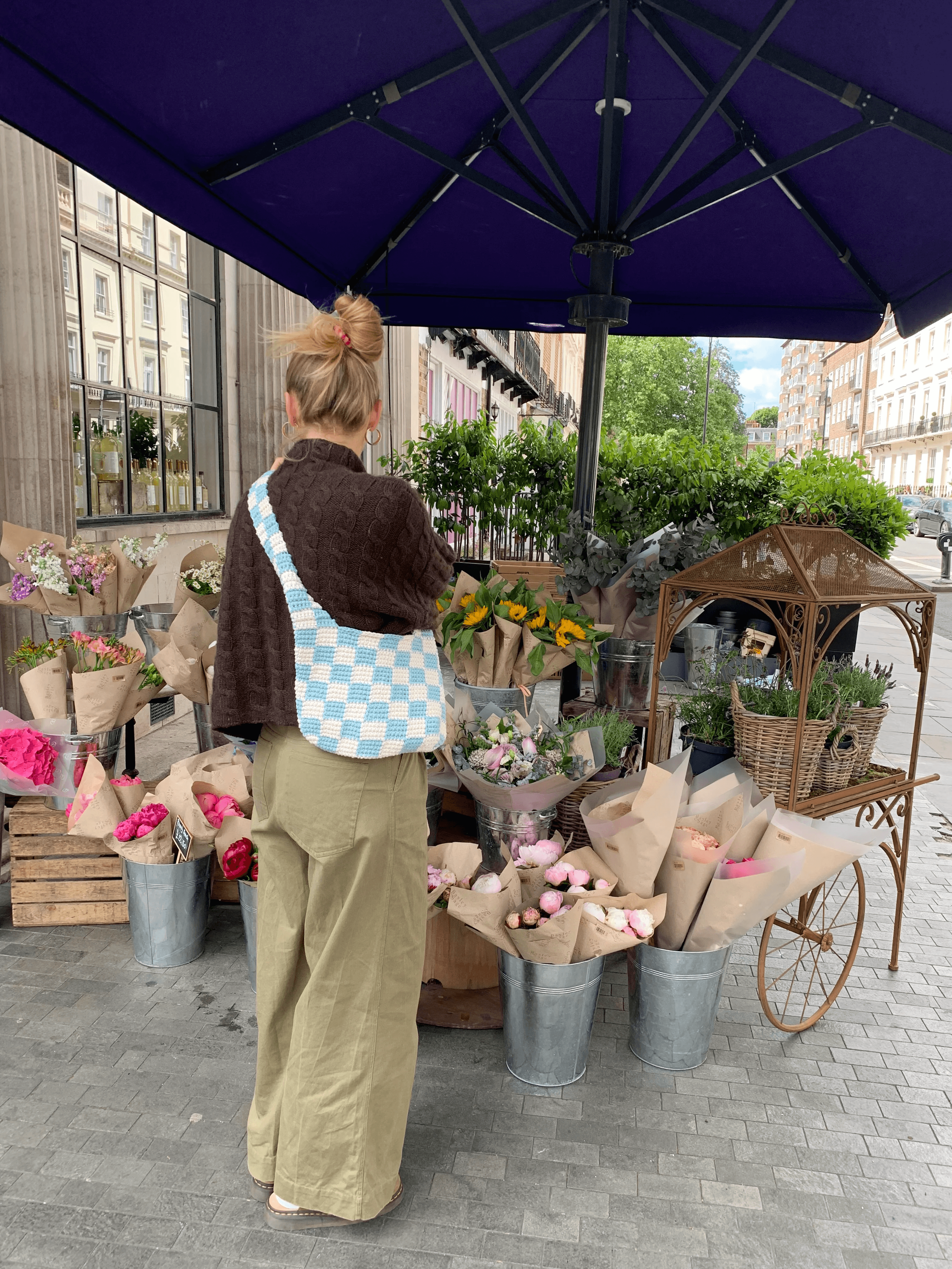A woman with a blue and white checkered bag stands under a large purple umbrella, browsing a vibrant assortment of fresh flowers wrapped in brown paper at a charming outdoor flower stall with an elegant green cart nearby, set on a cobblestone street in a city.