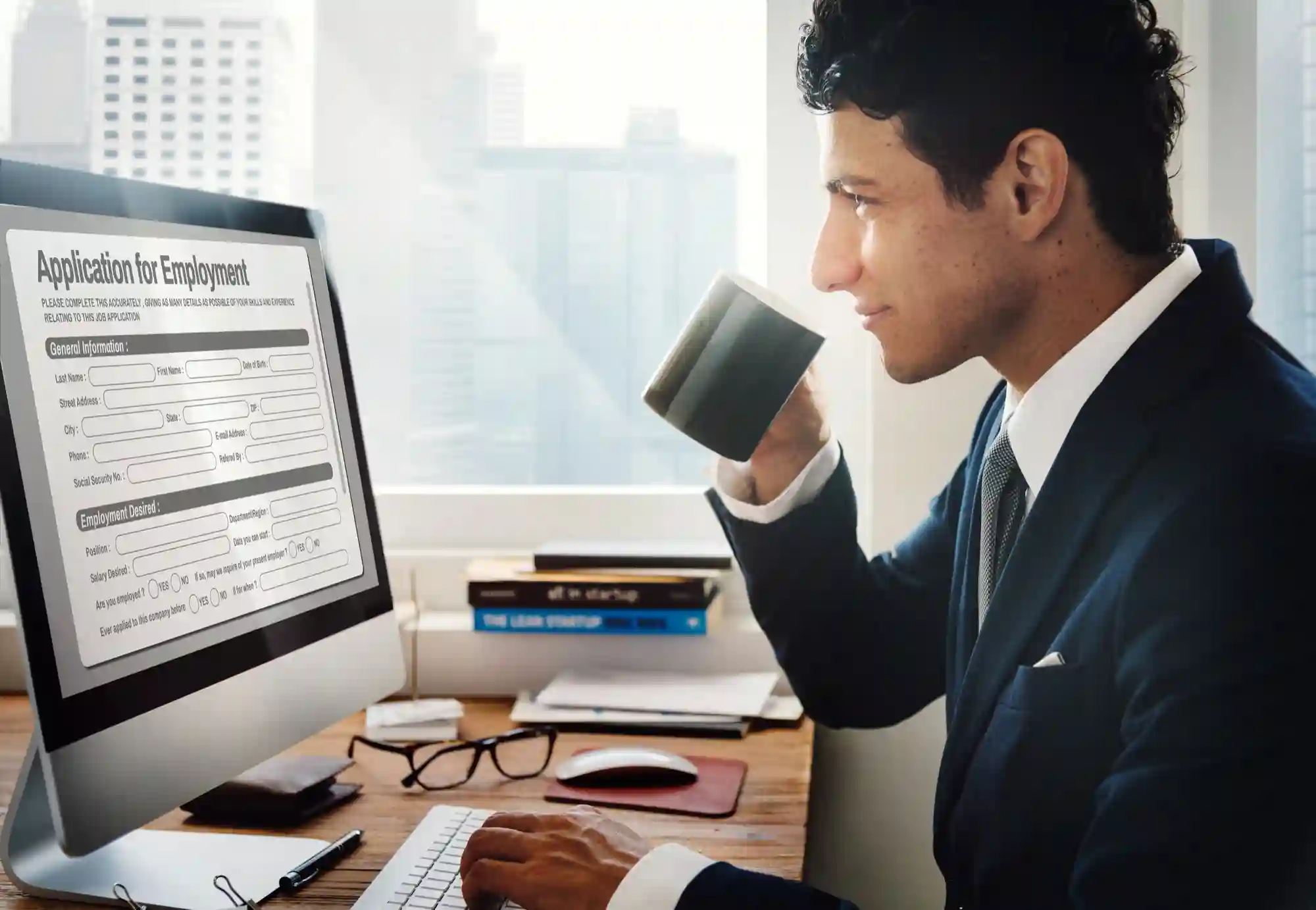A professional man in a suit drinks coffee while filling out an online employment application on his desktop computer.