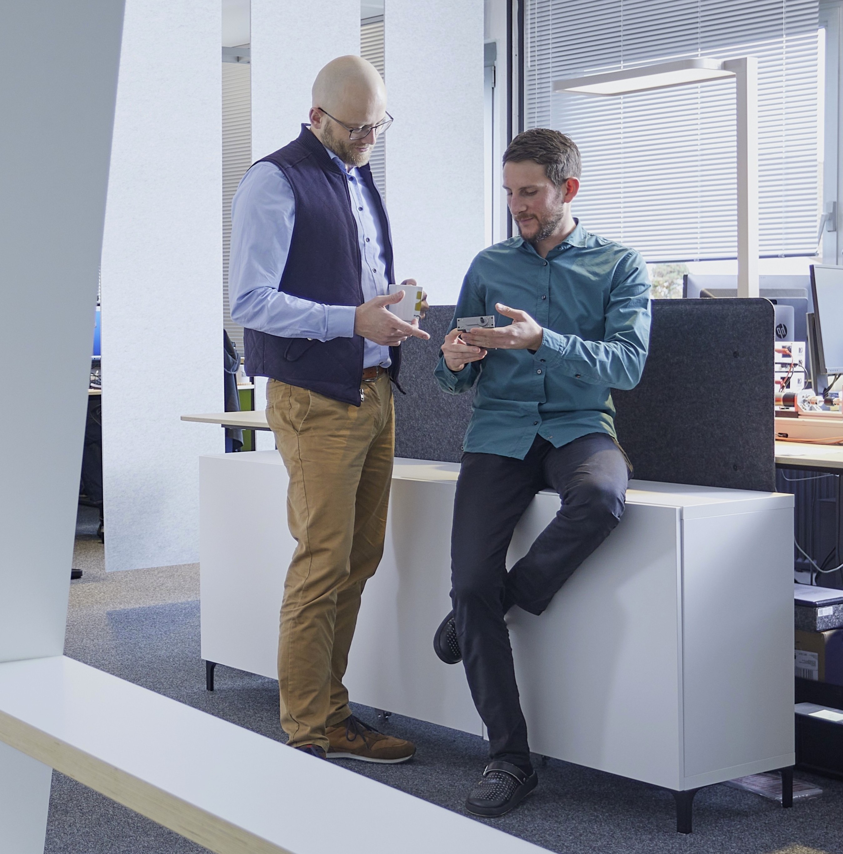Two colleagues in an office discussing something on a device. One man sits on a credenza holding it, while the other stands with a coffee mug and points at the screen.