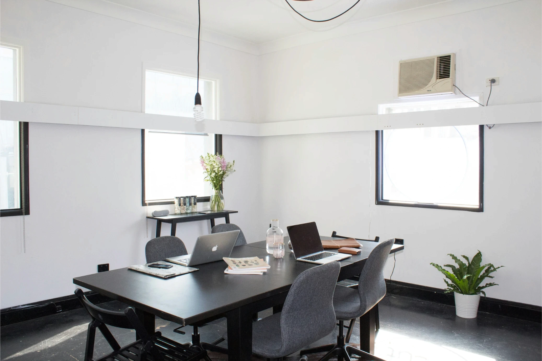 Bright, minimalist office space with a black table, gray chairs, and laptops. White walls, natural light, potted plants, and hanging bulb create a calm vibe.