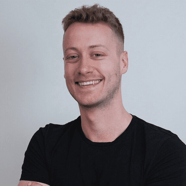 Professional headshot of a smiling young man with short blonde hair wearing a black t-shirt against a plain white background.