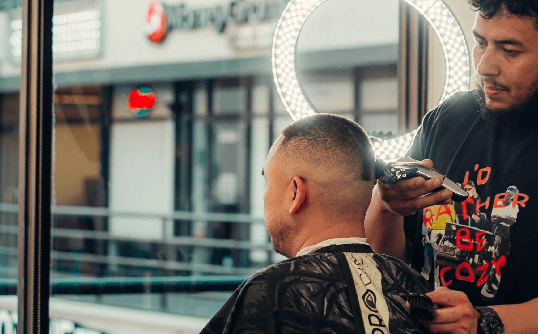 A barber shapes a client’s fade haircut using clippers near a bright ring light in a modern barbershop.